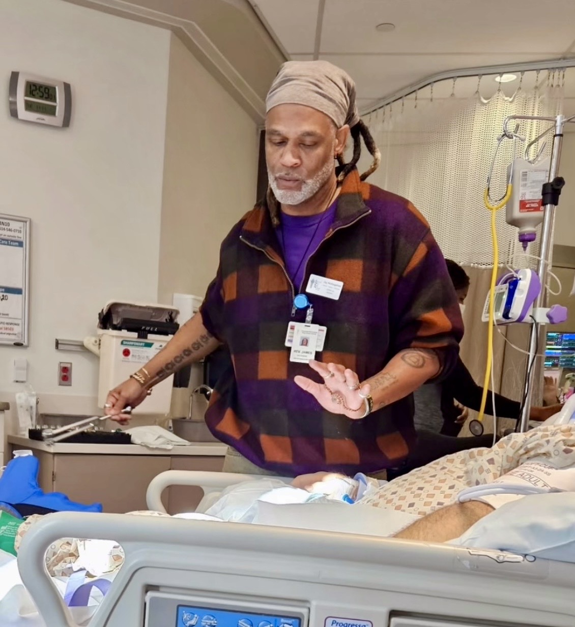 Medical professional with a head covering and plaid shirt standing beside a patient in a hospital bed, medical equipment visible.