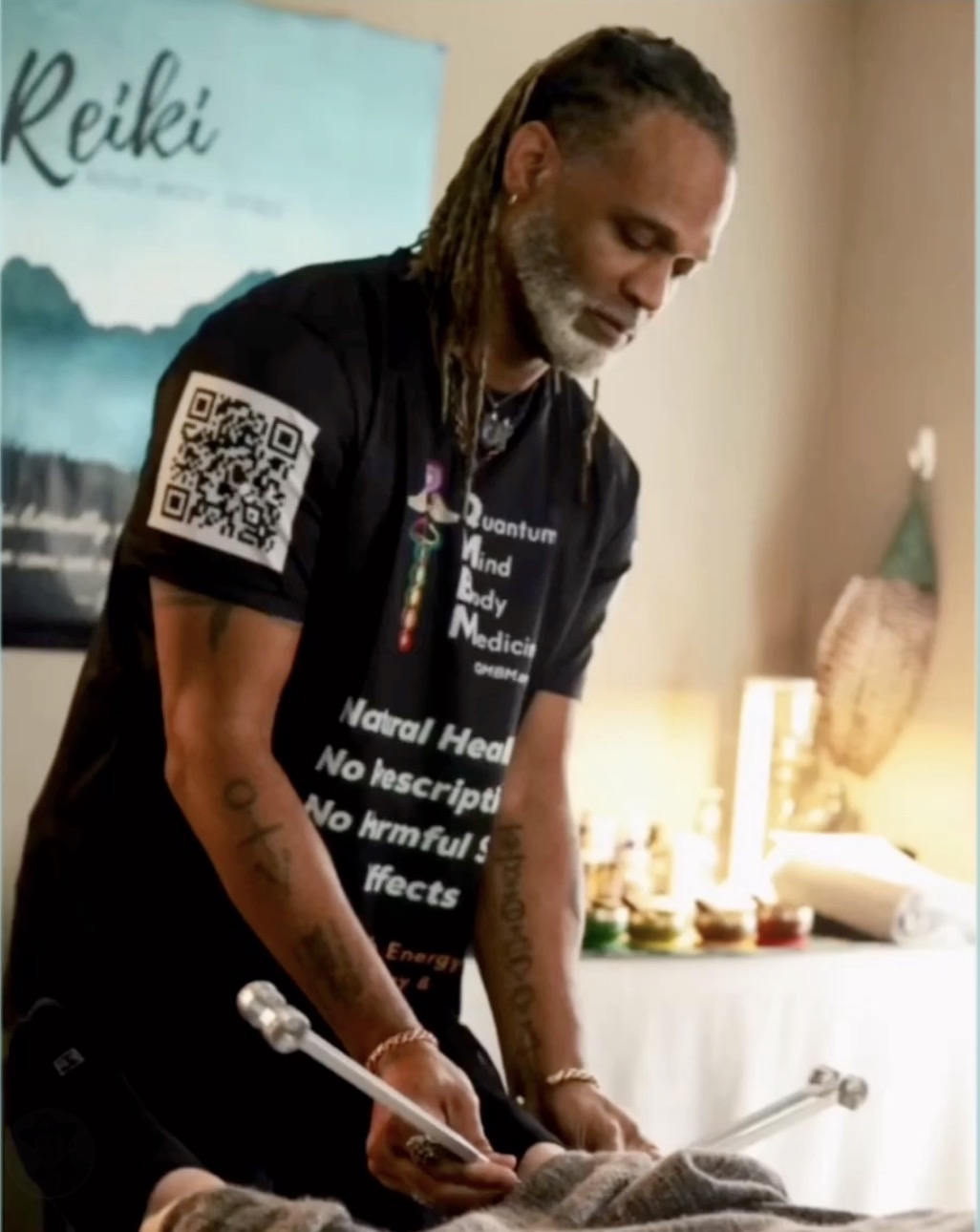 Man with dreadlocks and gray beard using tuning forks in a room with a Reiki sign on the wall.