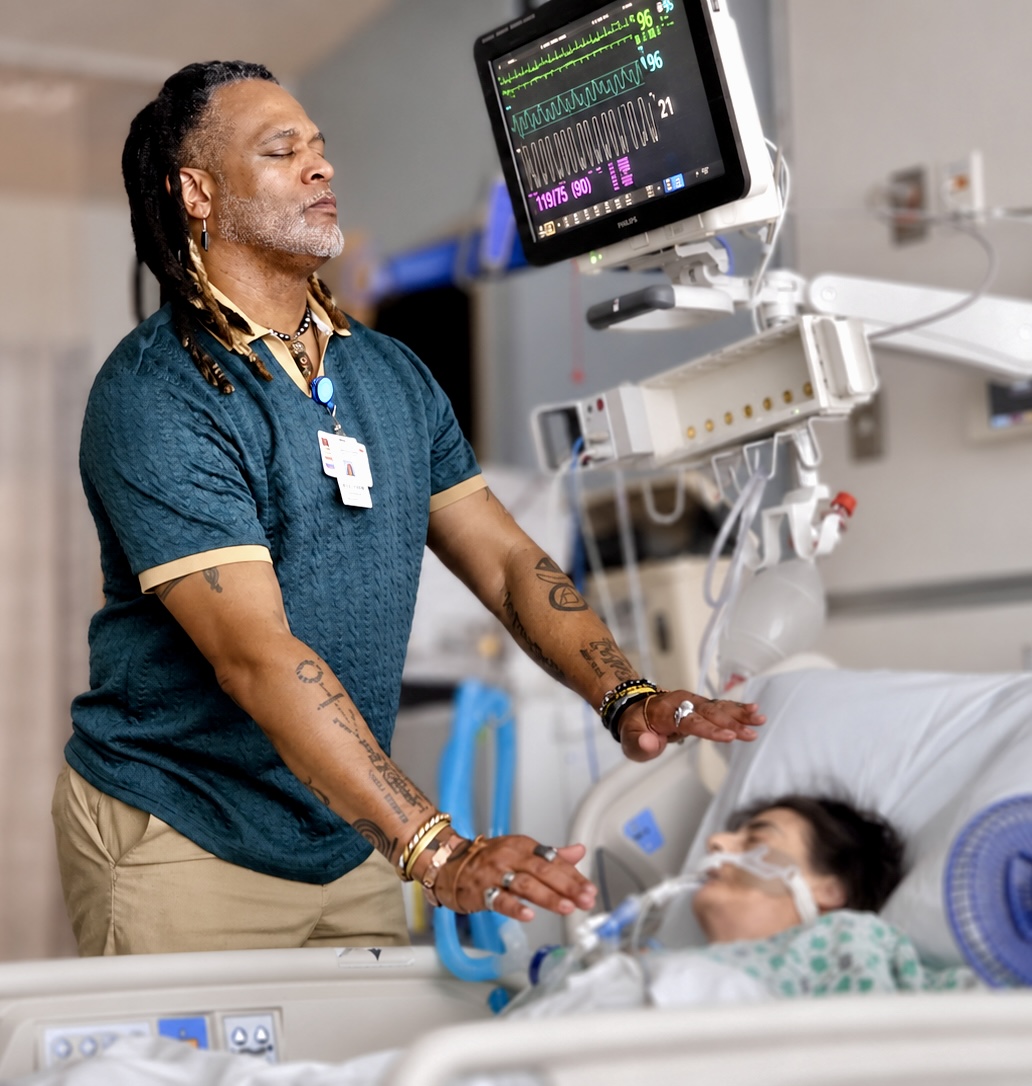A man with dreadlocks and tattoos stands beside a hospital bed, monitoring a patient connected to medical equipment.