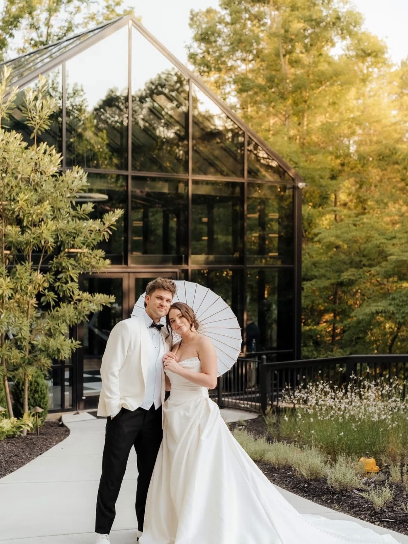 A couple in wedding attire standing outside a glass building, holding a white umbrella, surrounded by trees and greenery.