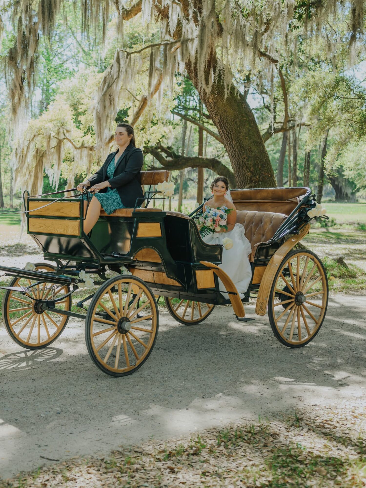 Two women sit in a vintage horse-drawn carriage outdoors under a large tree with hanging moss, surrounded by greenery.