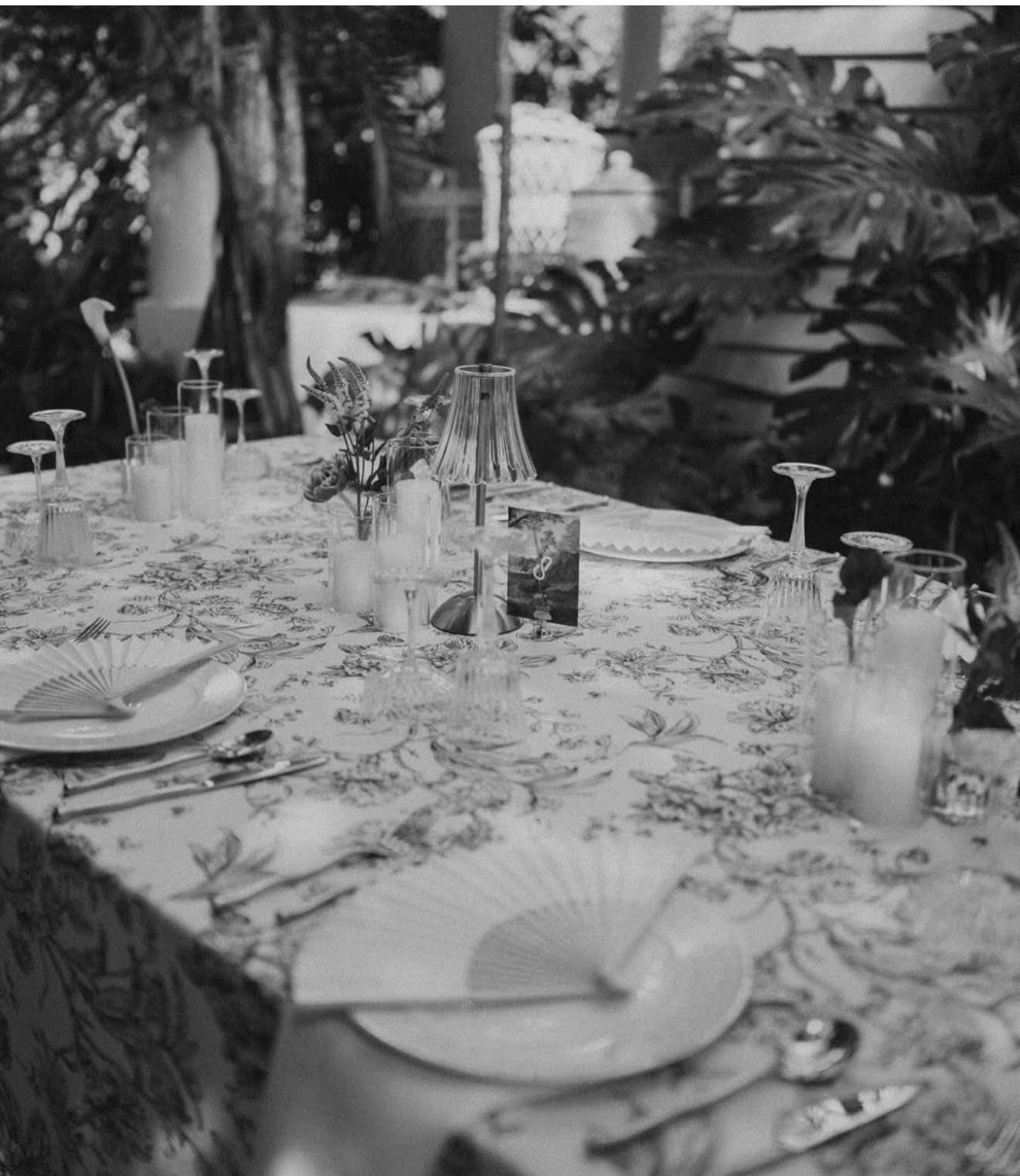 Table set with plates, glasses, a small lamp, and floral centerpiece outdoors, surrounded by plants and trees.
