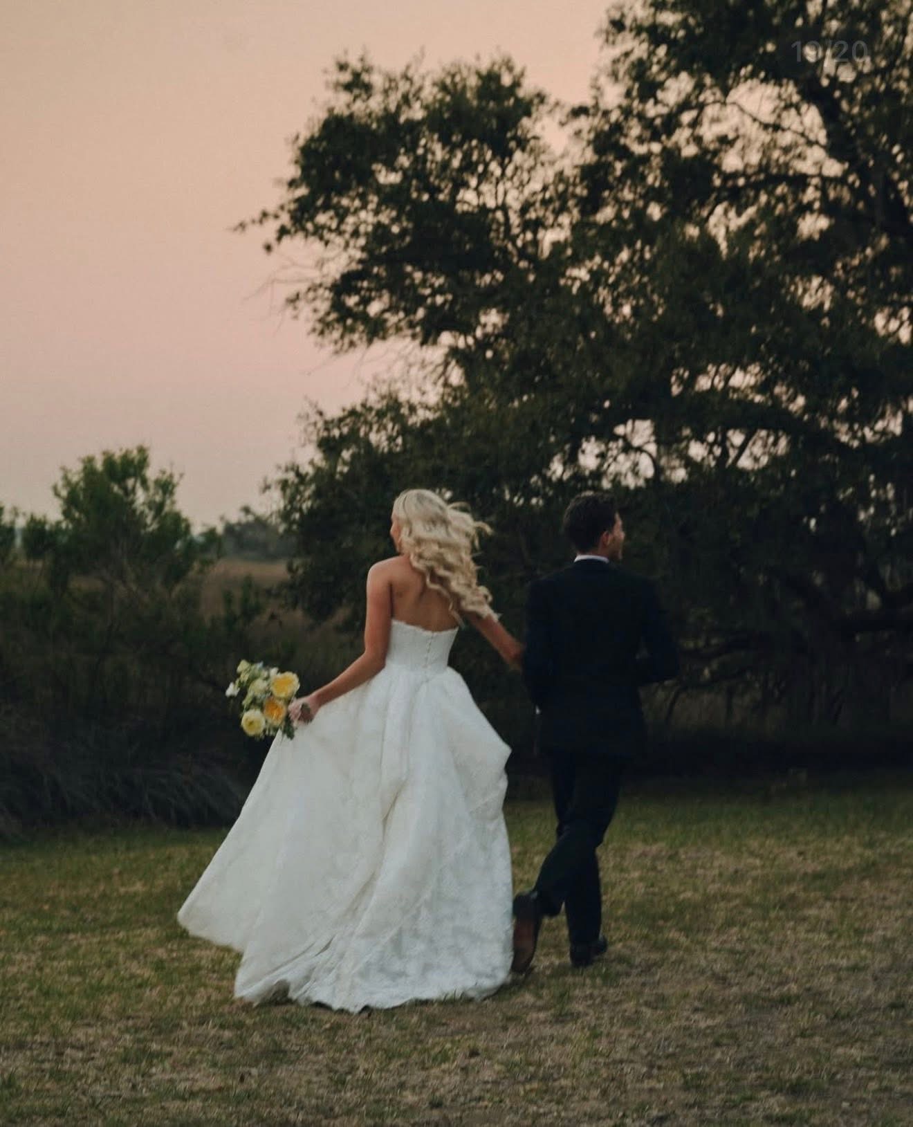 Couple walking outdoors, woman in wedding dress holding flowers, man in suit, large tree in background