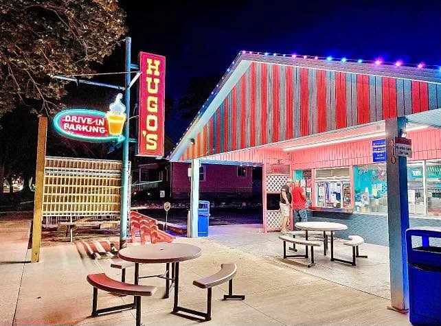 Night scene of a drive-in burger restaurant with outdoor seating, illuminated signage, and a striped roof, empty tables and a person at the counter.