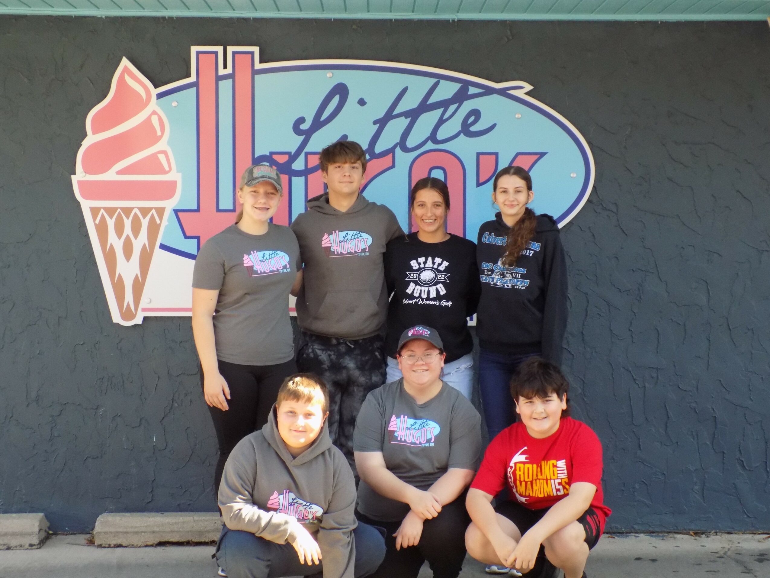 Group of six children standing and sitting in front of an ice cream shop sign, smiling, with a dark wall background.