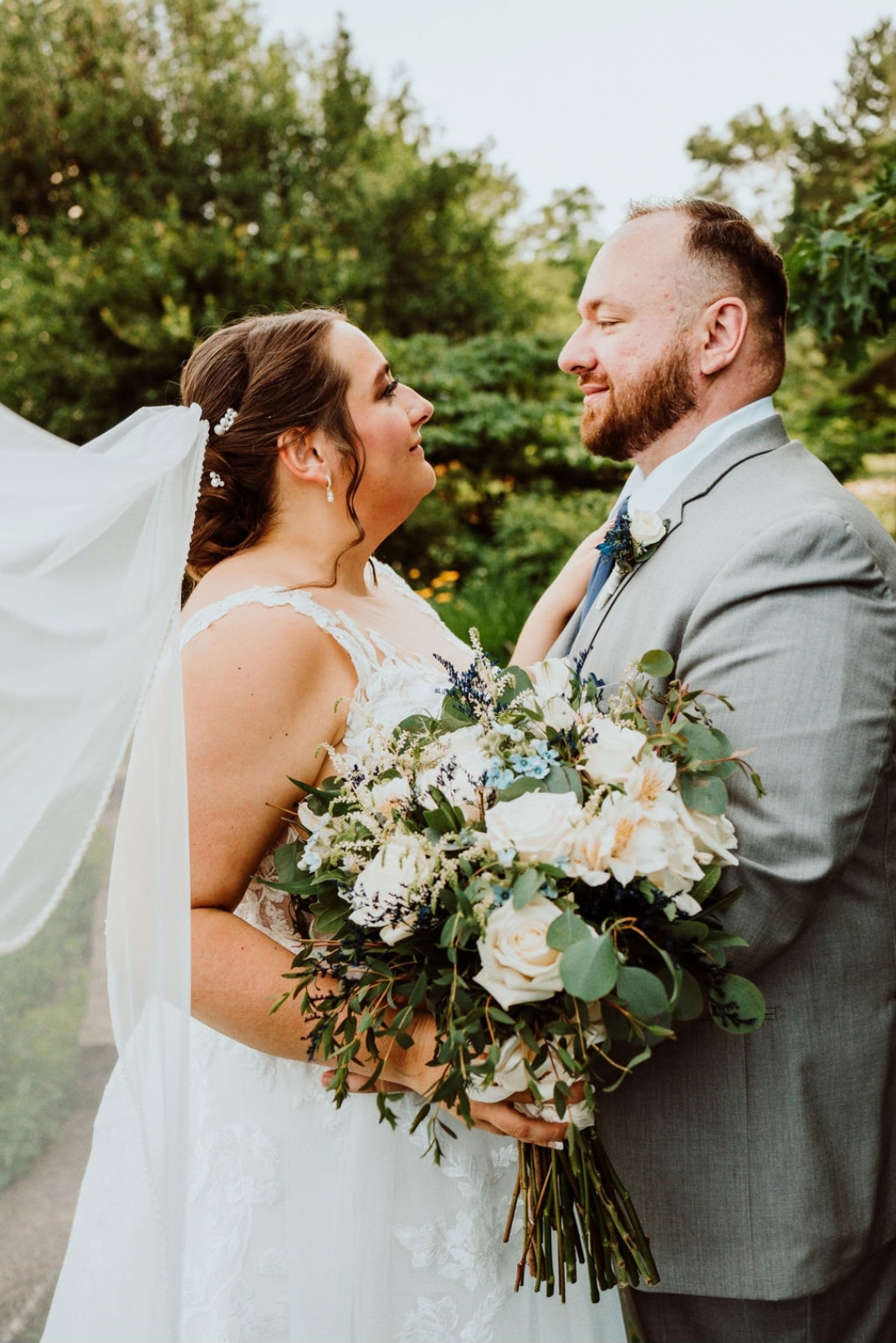 Bride and groom face each other outdoors, smiling, with the bride holding a large bouquet of flowers, greenery, and white blooms.