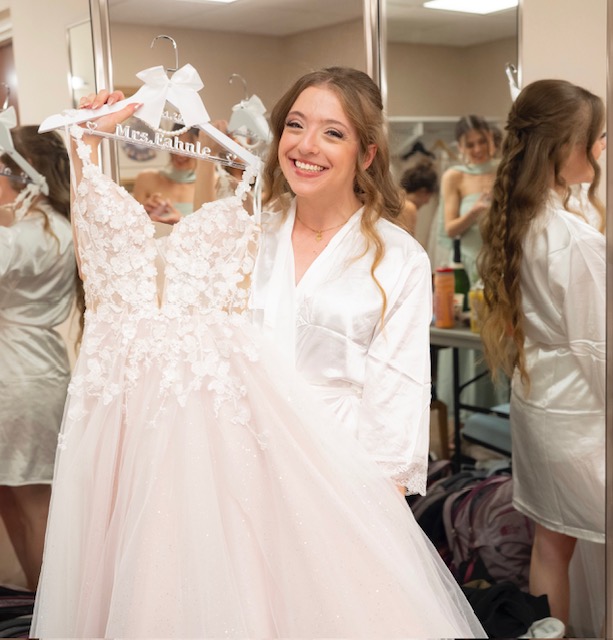 Smiling woman holding a wedding dress on a hanger in a dressing room with mirrors and other people. 20 words.