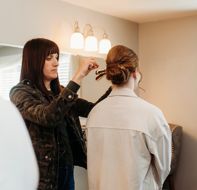 A woman with dark hair styles a person's hair in a room with a mirror and warm lighting.