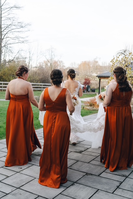 Four women in matching rust-colored dresses walk outdoors on a paved path, with trees and a building in the background.