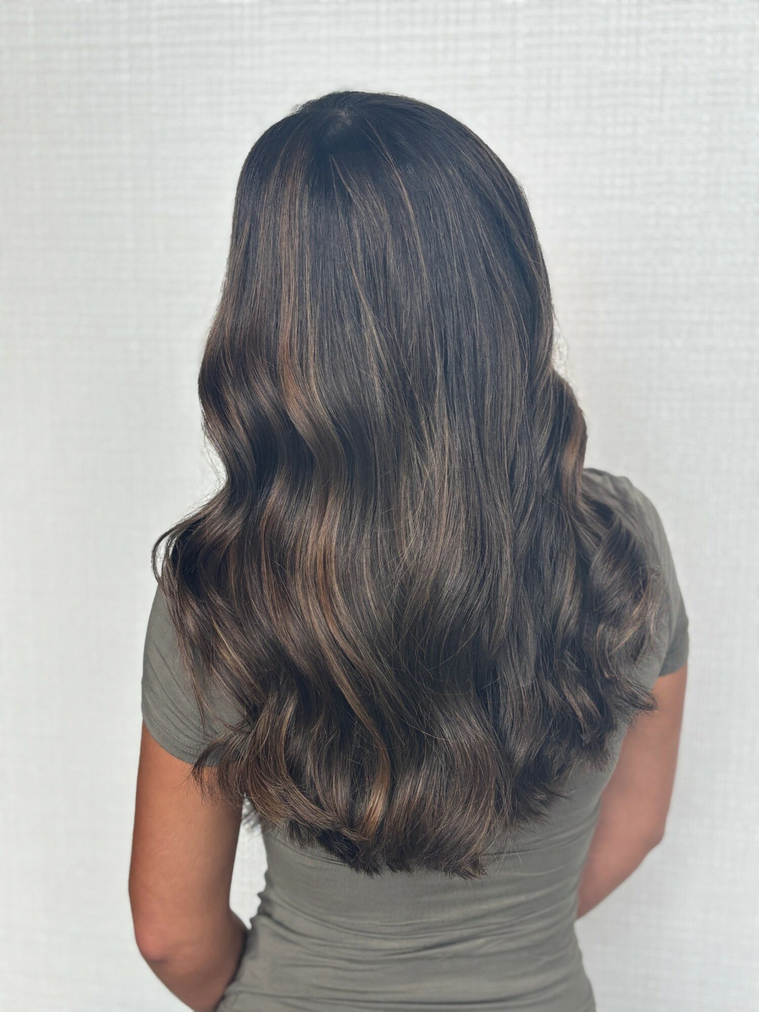 Back view of a woman with long, wavy brown hair wearing a gray top, standing against a plain background.