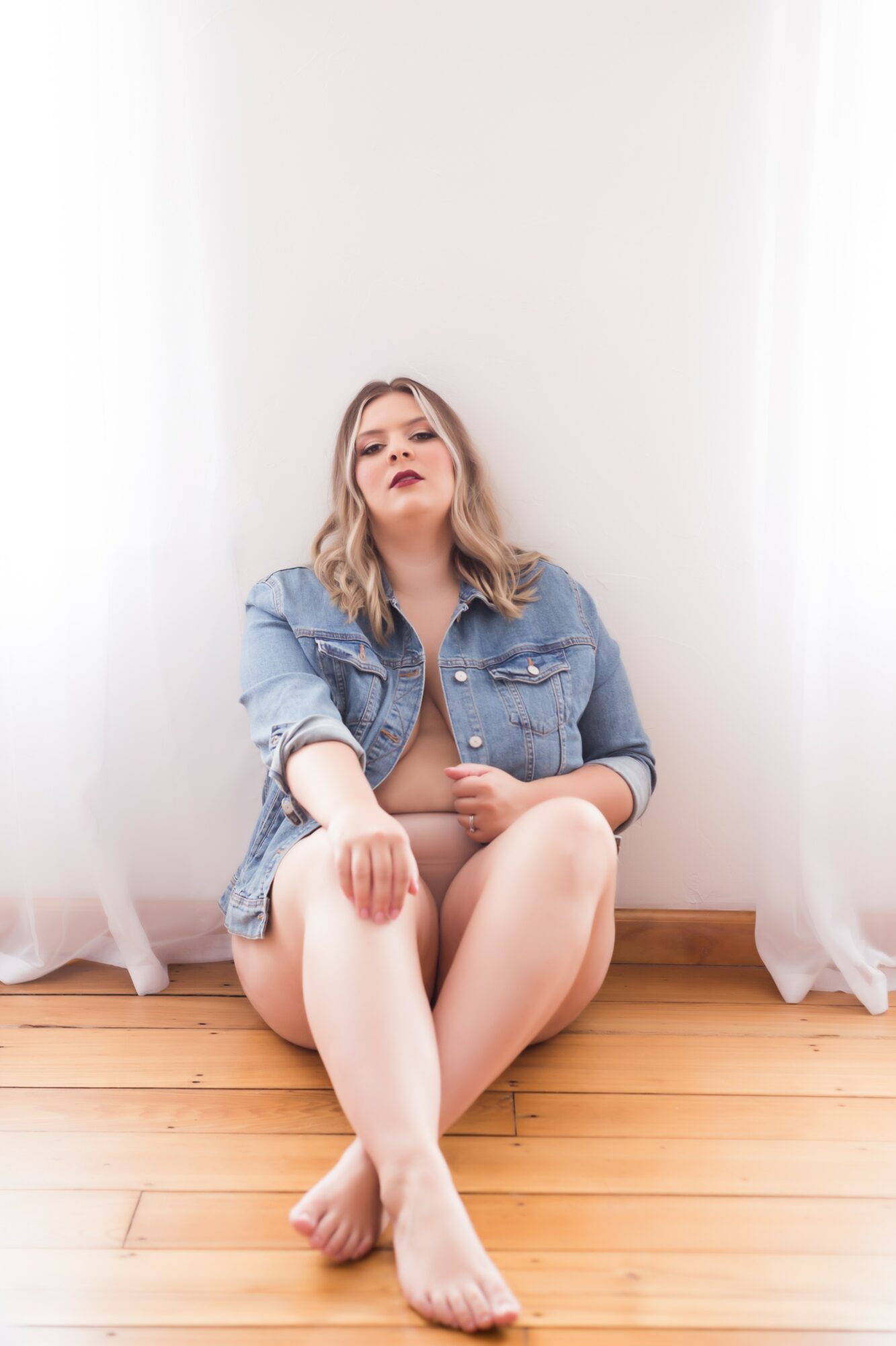 Young woman with blonde hair sitting on wooden floor, wearing denim jacket, against white wall and curtains.