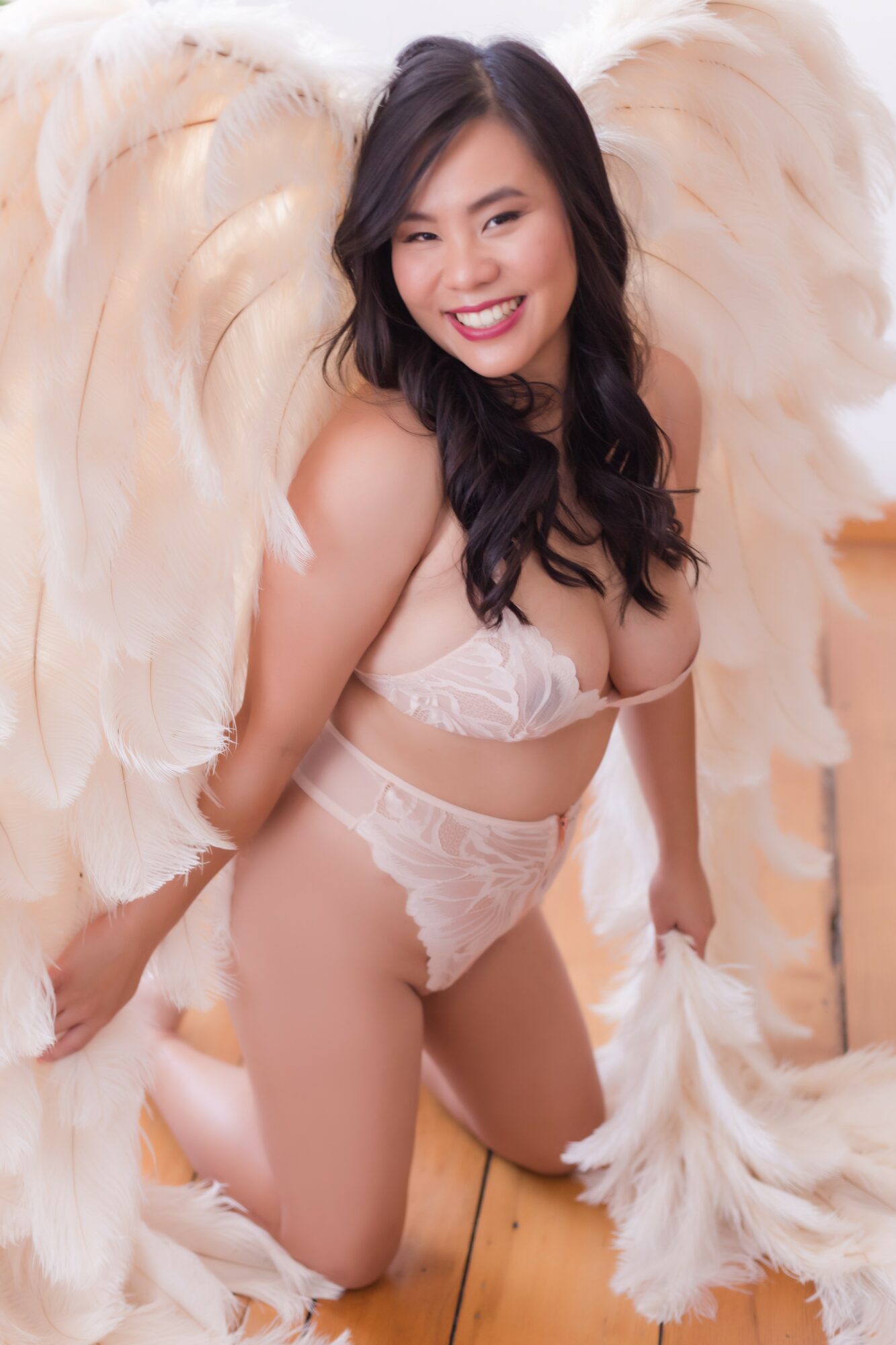 Young woman with large feathered wings, smiling, wearing lingerie, kneeling on a wooden floor, looking at the camera.
