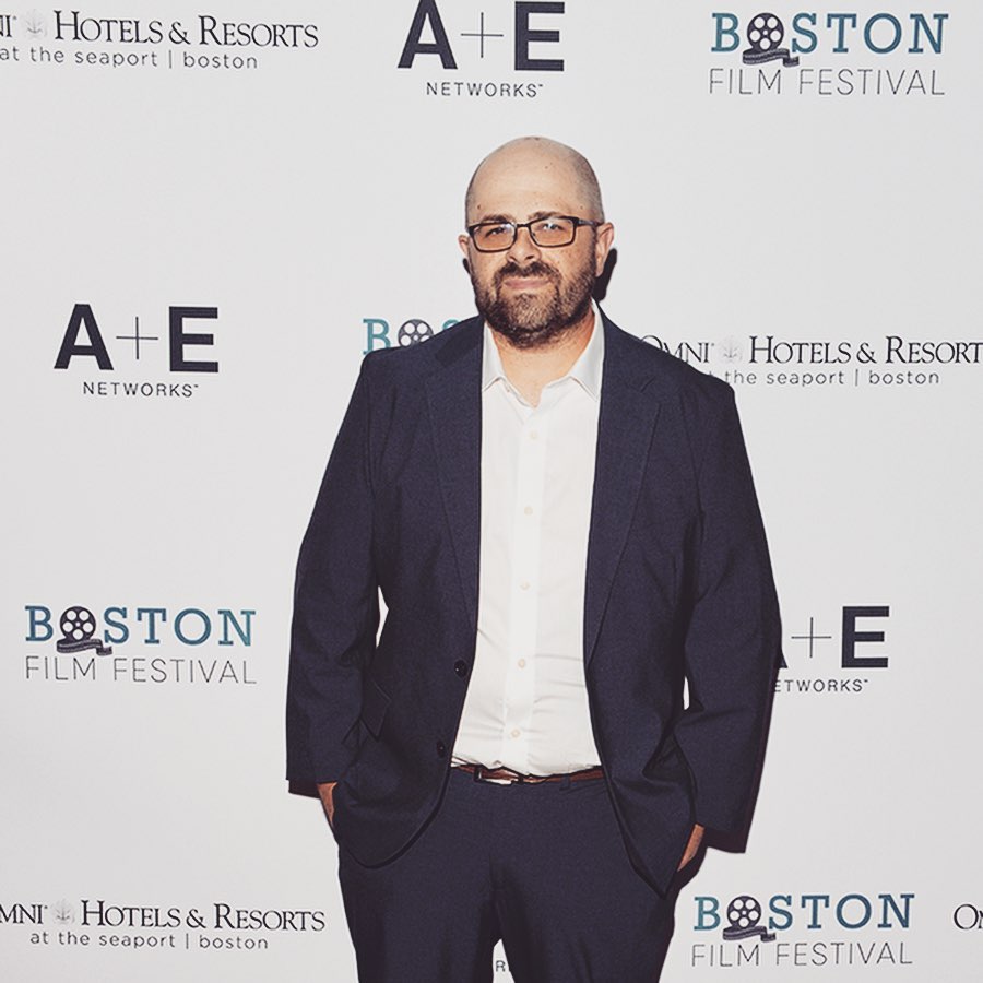 Man with glasses and beard in a suit standing in front of a white backdrop with logos and text.