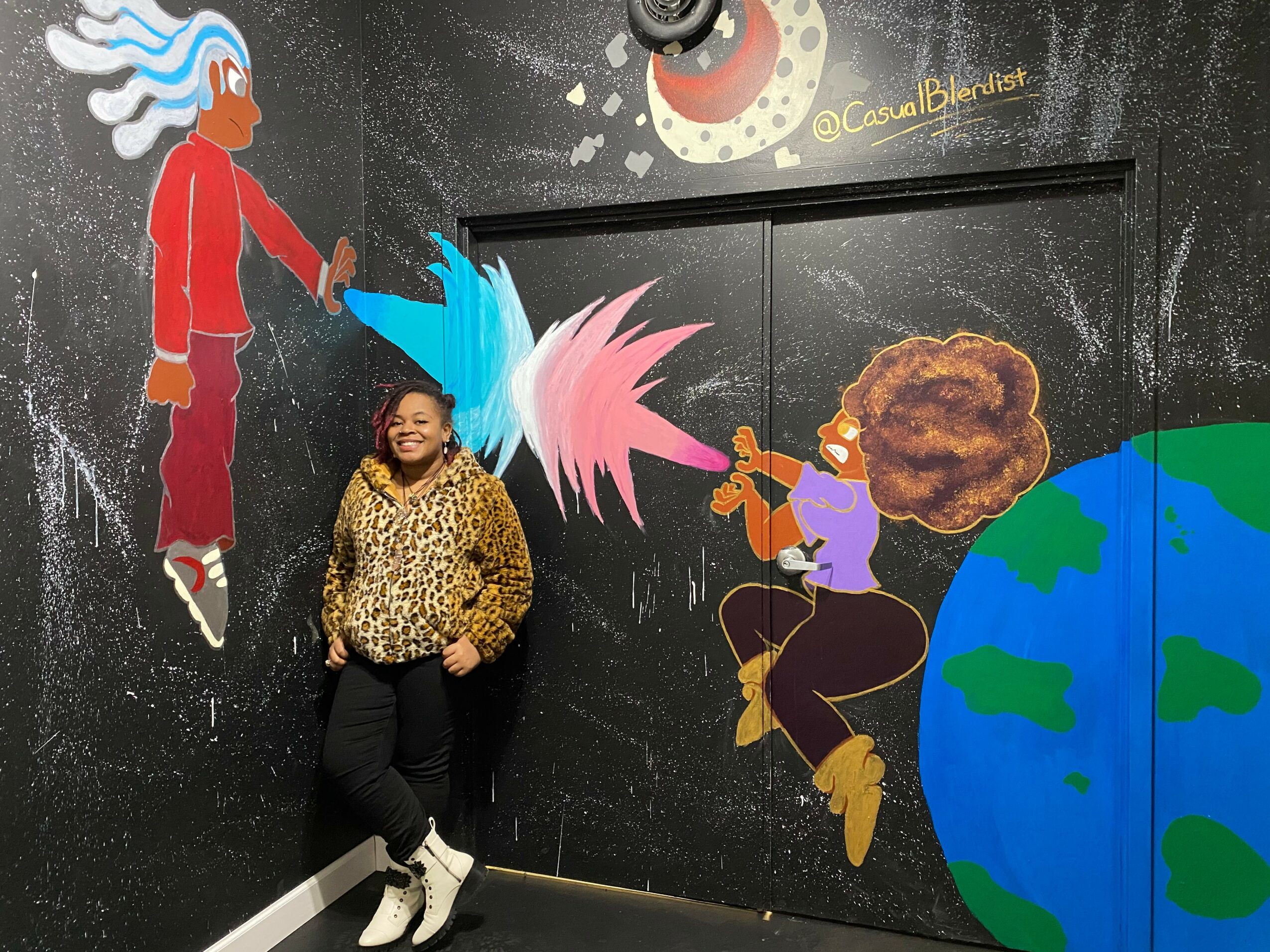 Woman standing in front of a colorful mural with a person flying, a musician, and a globe, smiling and posing.