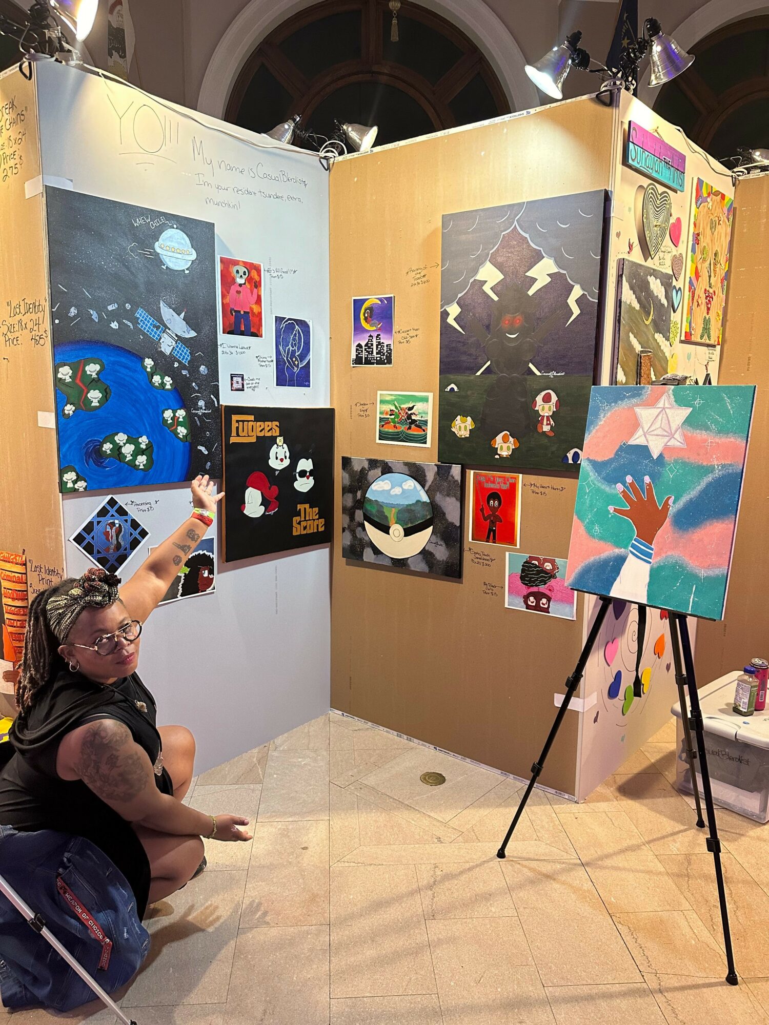 Display of colorful artwork and posters on a board, with a person sitting nearby, in an indoor setting.