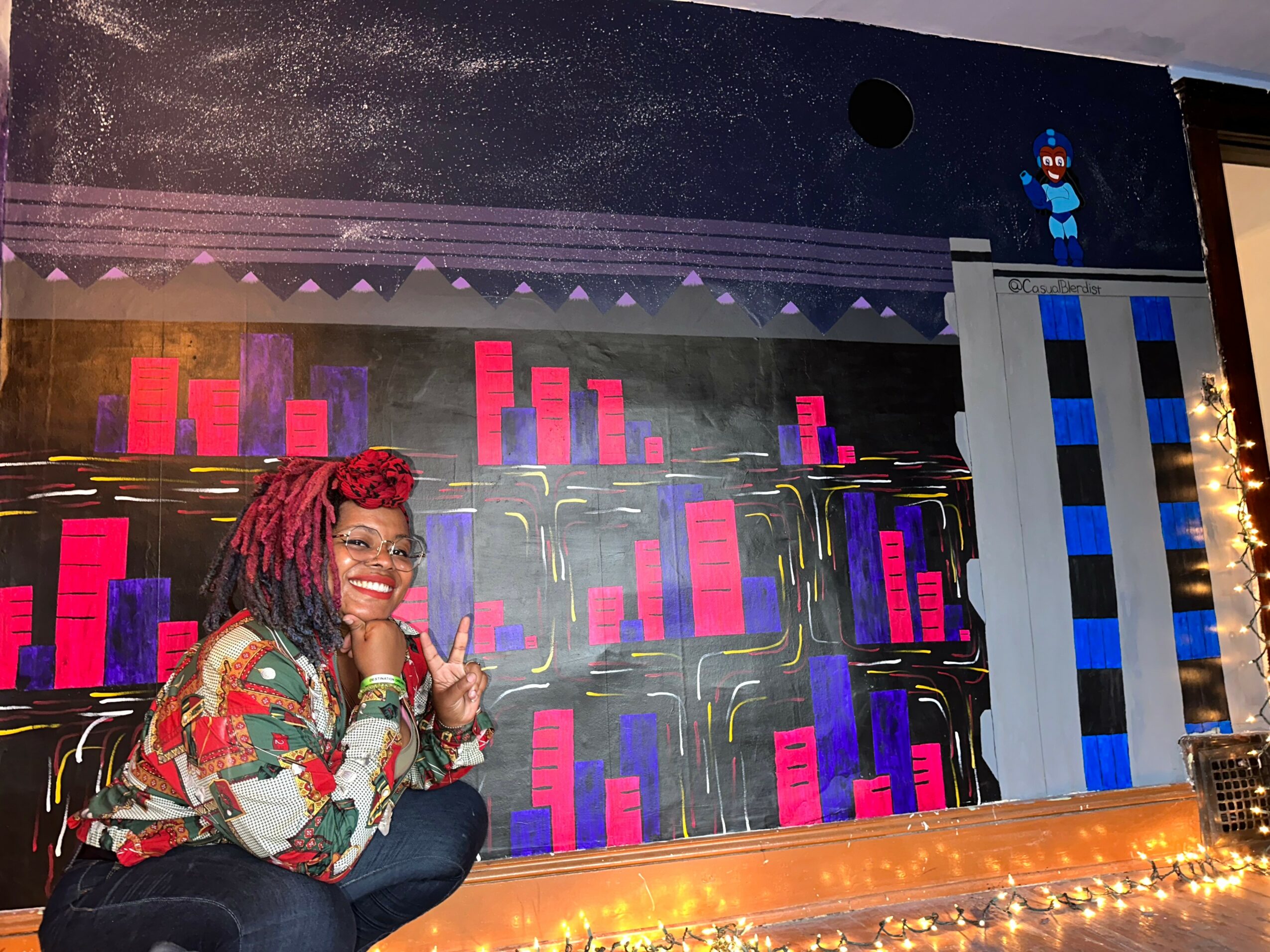 Person with red hair and glasses smiling, sitting in front of a colorful cityscape backdrop with pink and purple buildings, and a small figure on a ledge above.