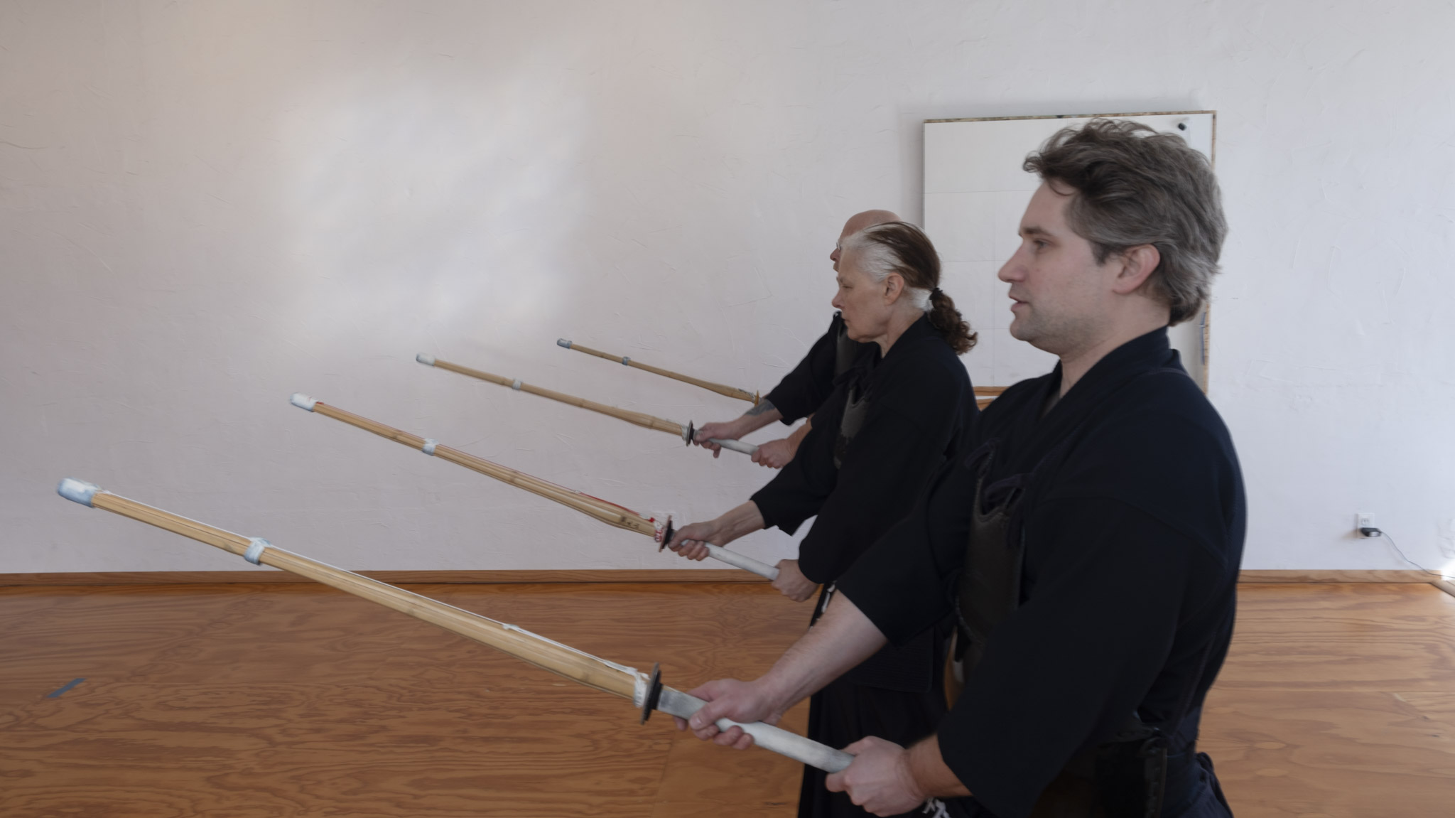 Three people stand in a line holding long wooden sticks, facing a blank white wall, in a room with wooden flooring.