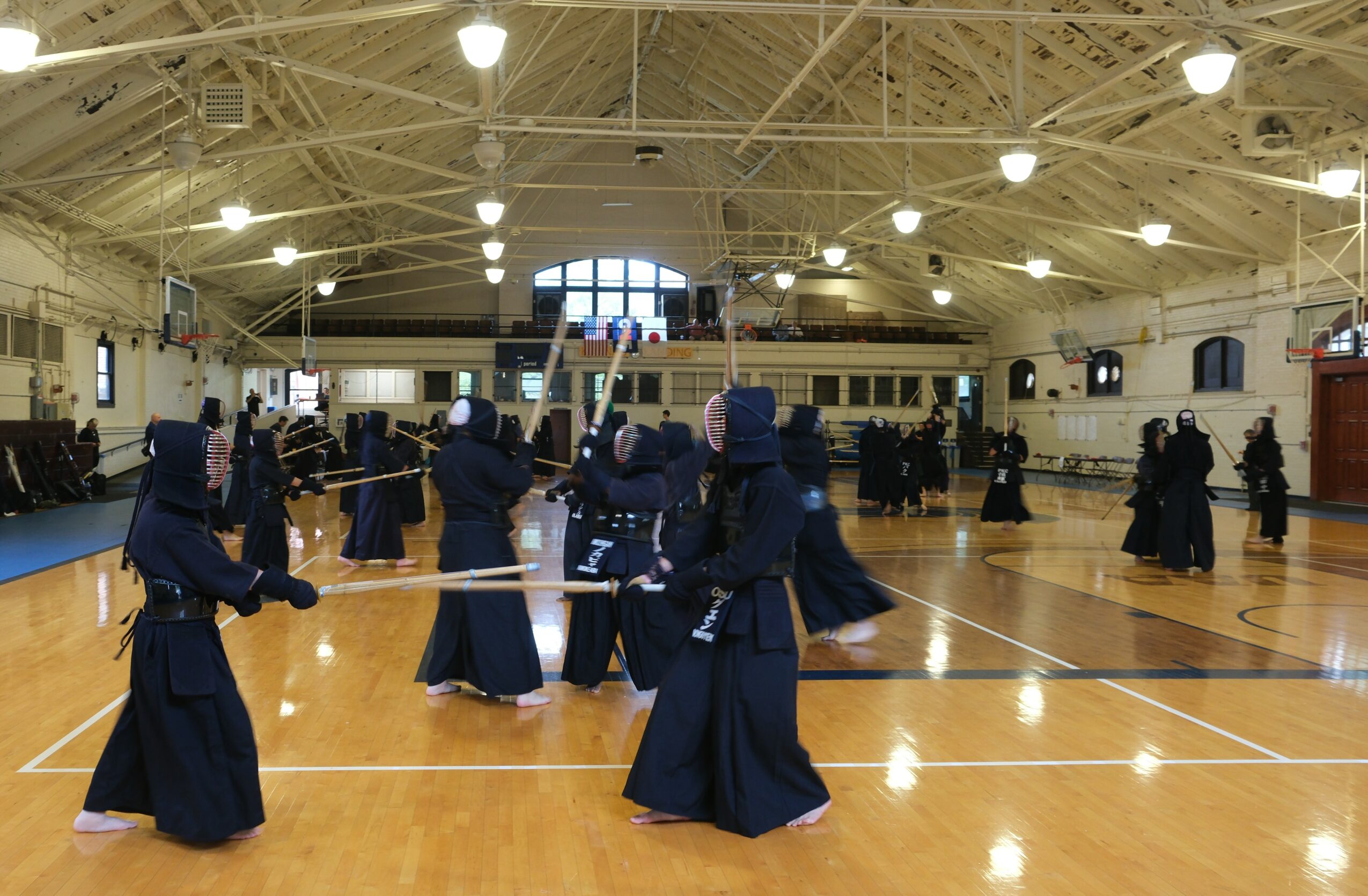 Group of people practicing martial arts in a gymnasium with high ceiling and bright lighting.
