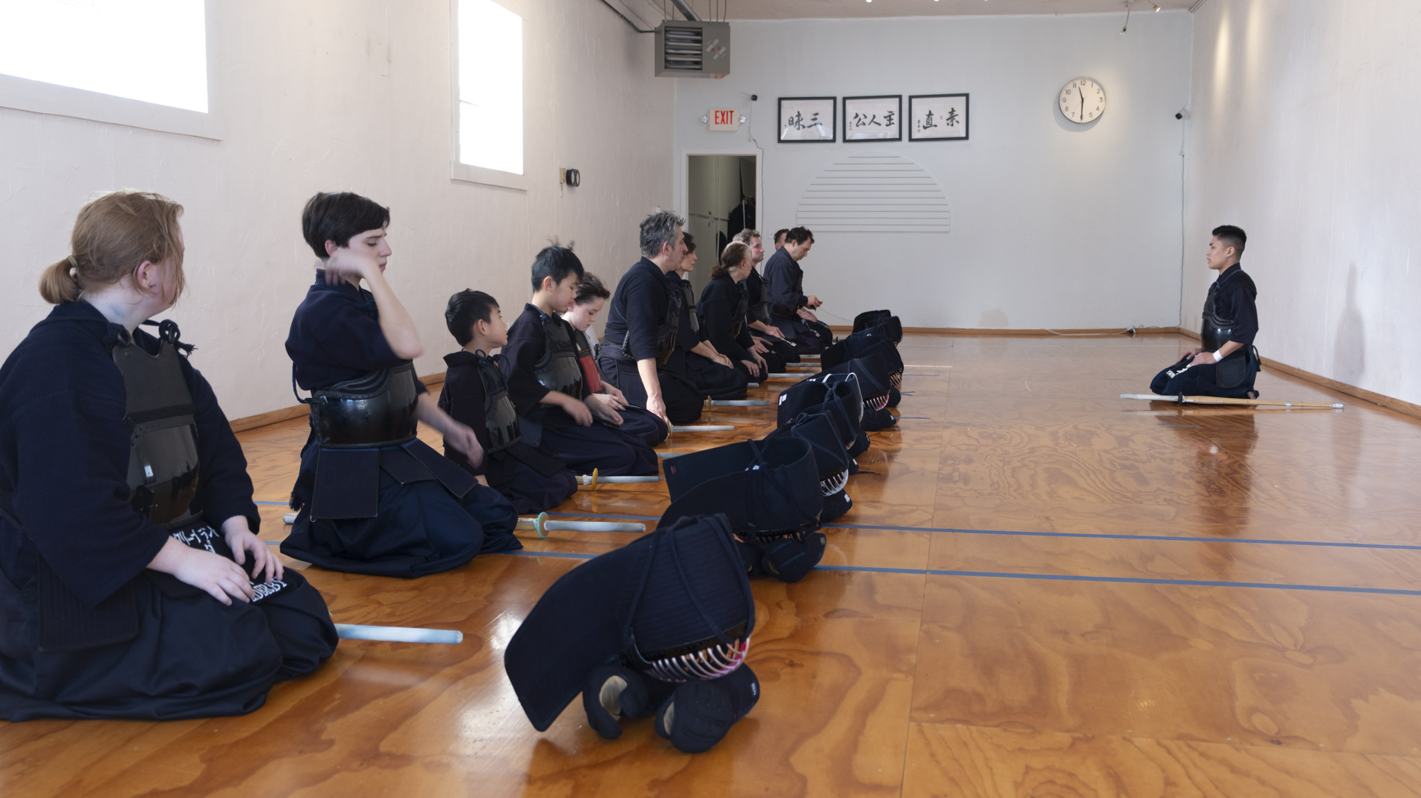 Group of children in uniform kneeling and bowing in a spacious room with wooden floor, white walls, and framed pictures.