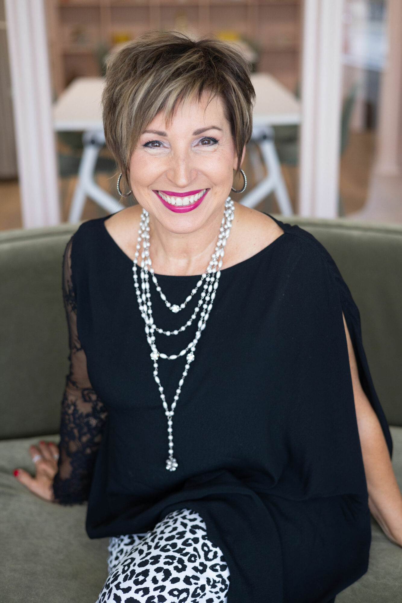 Woman with short hair, wearing a black top and multiple pearl necklaces, smiling, sitting on a sofa in a room.