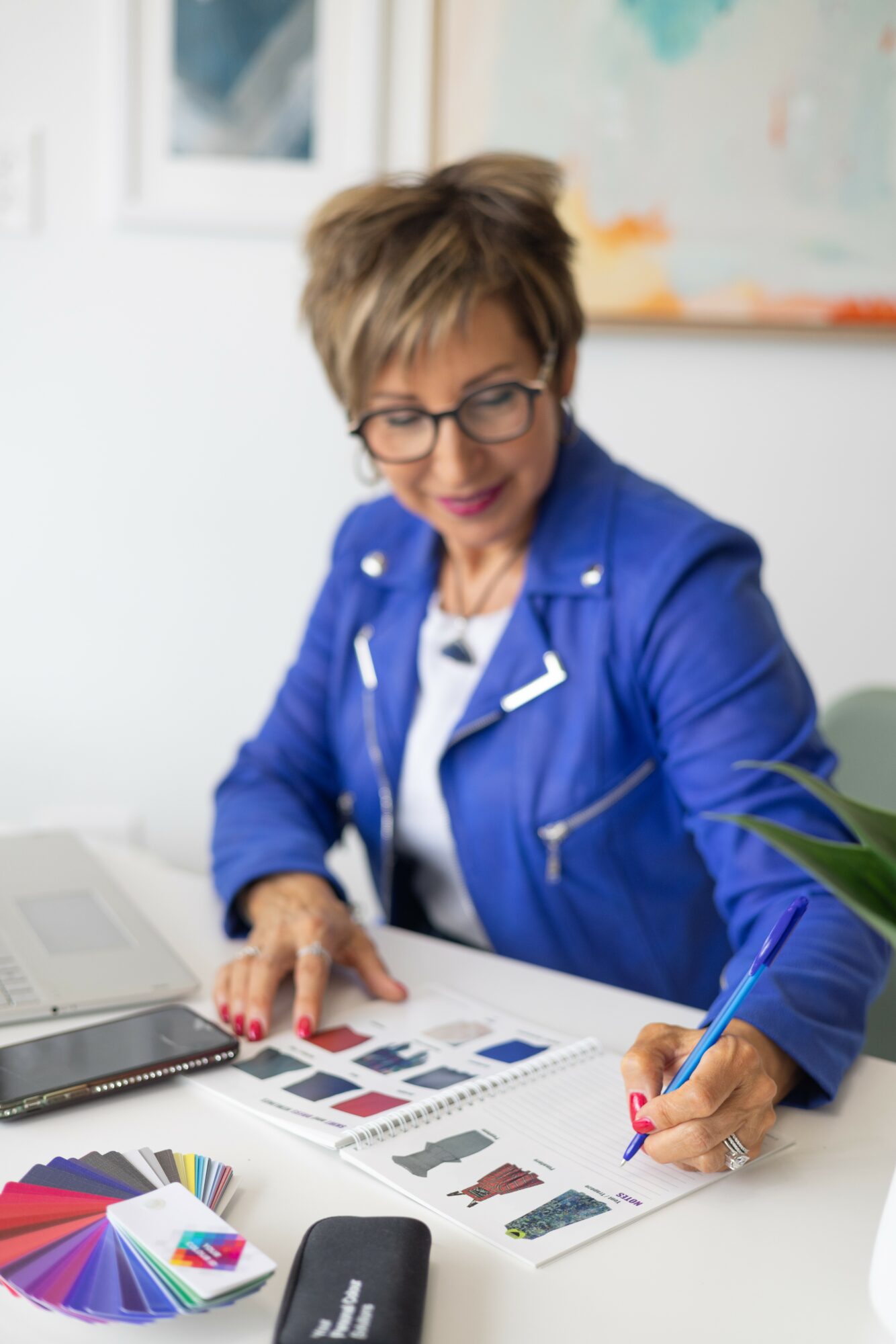 Woman with short hair and glasses in a blue jacket working at a desk with color samples and a pen.