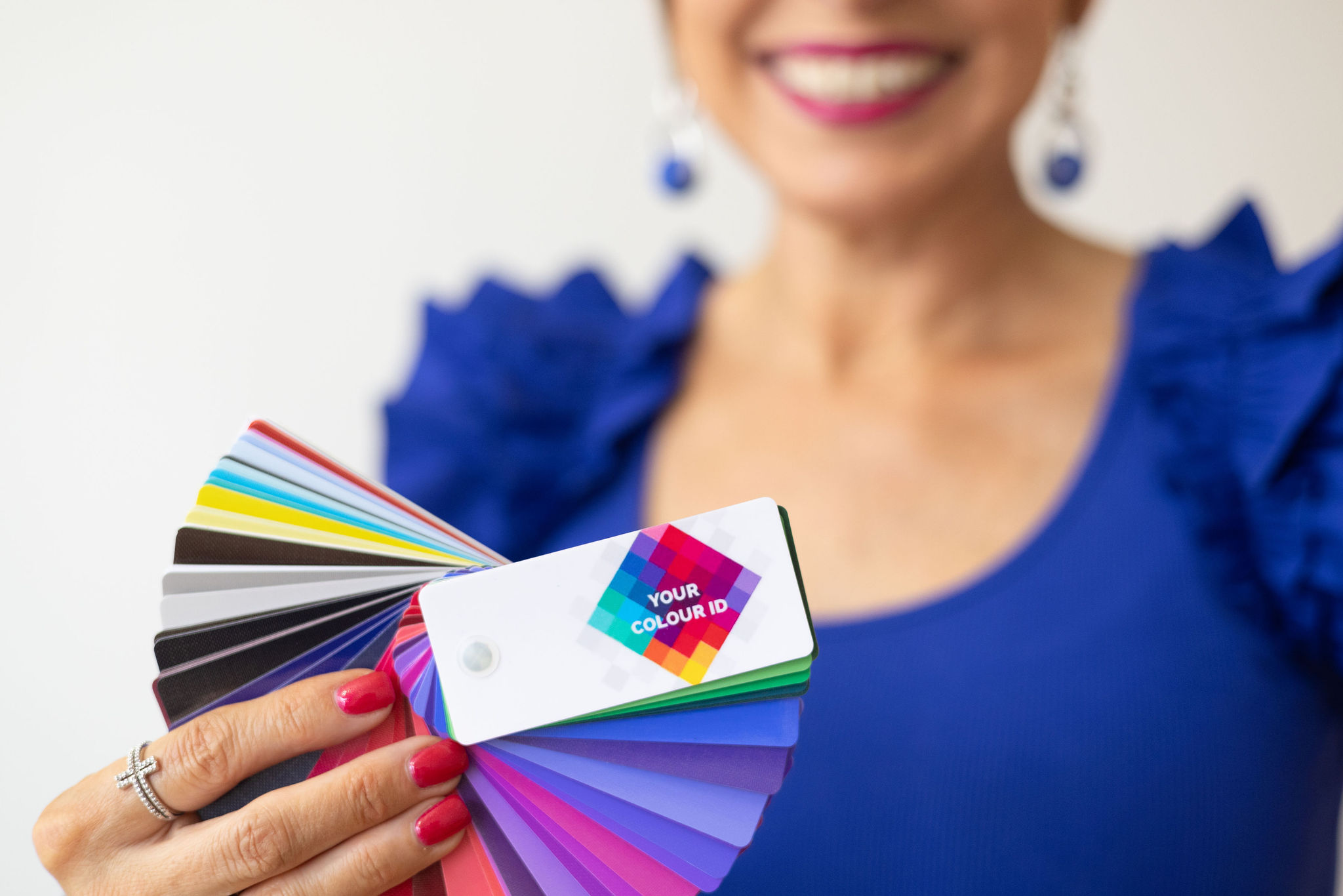 Person holding color swatches, wearing a blue top with ruffled shoulders, smiling, with pink nail polish and earrings.
