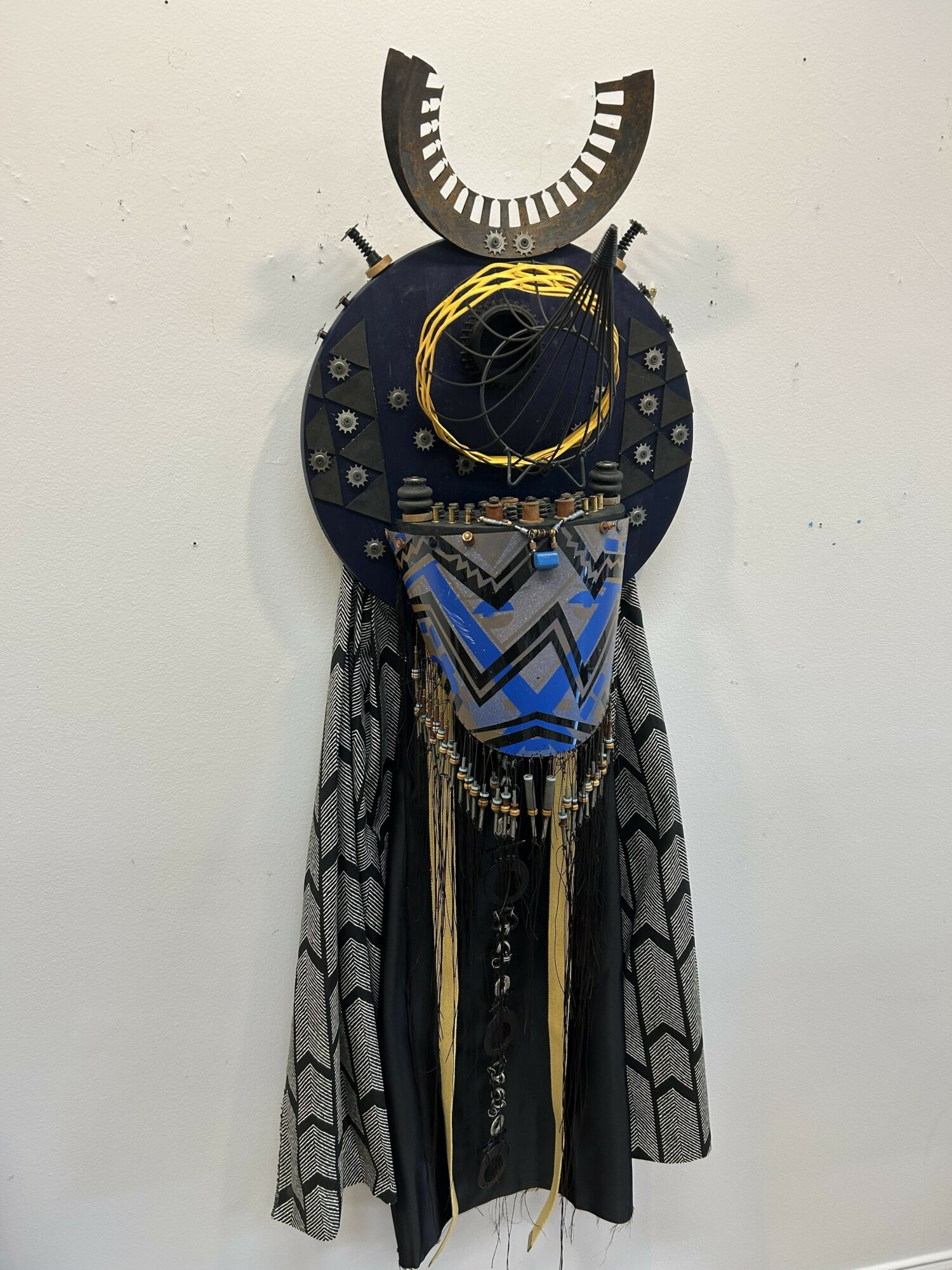 Decorative wall hanging with circular and geometric elements, fabric, and hanging accessories, resembling a tribal or artistic mask or sculpture.