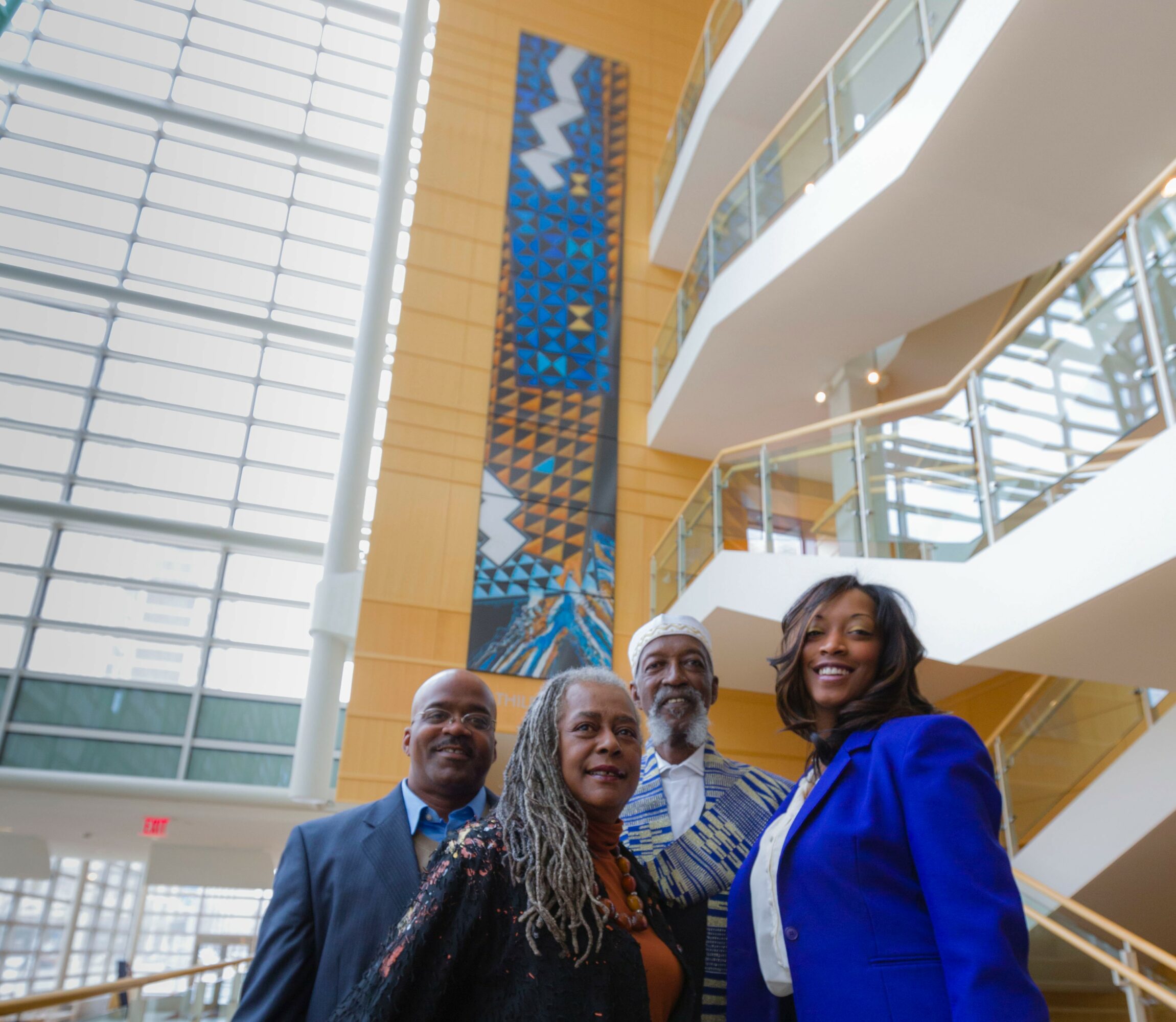 Four people standing inside a modern building with large windows and a colorful vertical artwork behind them.