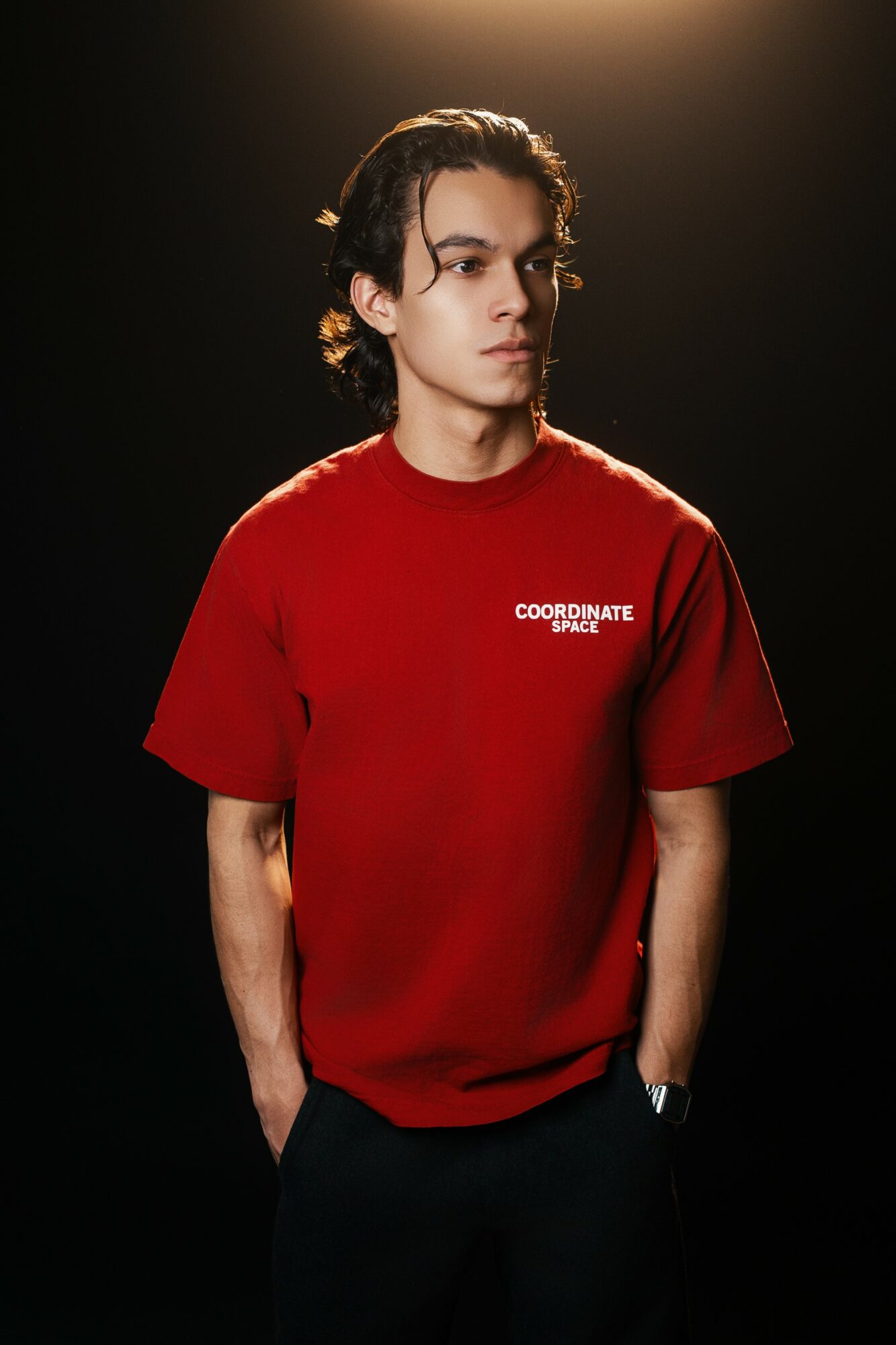 Young man with dark, wavy hair wearing a red T-shirt with white text, standing against a dark background.