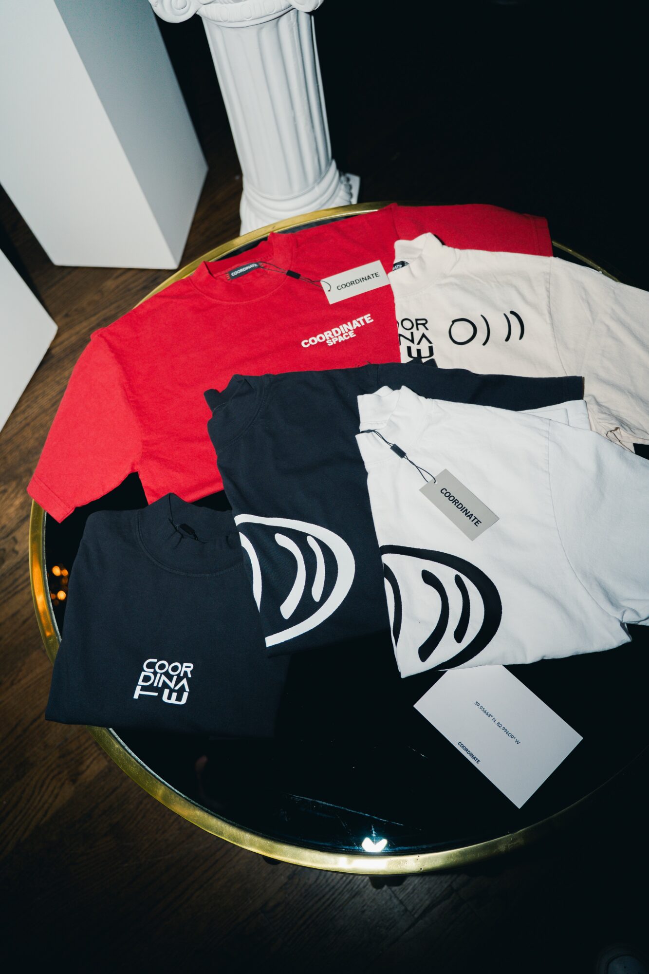 Multiple folded t-shirts in red, white, and black on a reflective black surface, with tags attached.