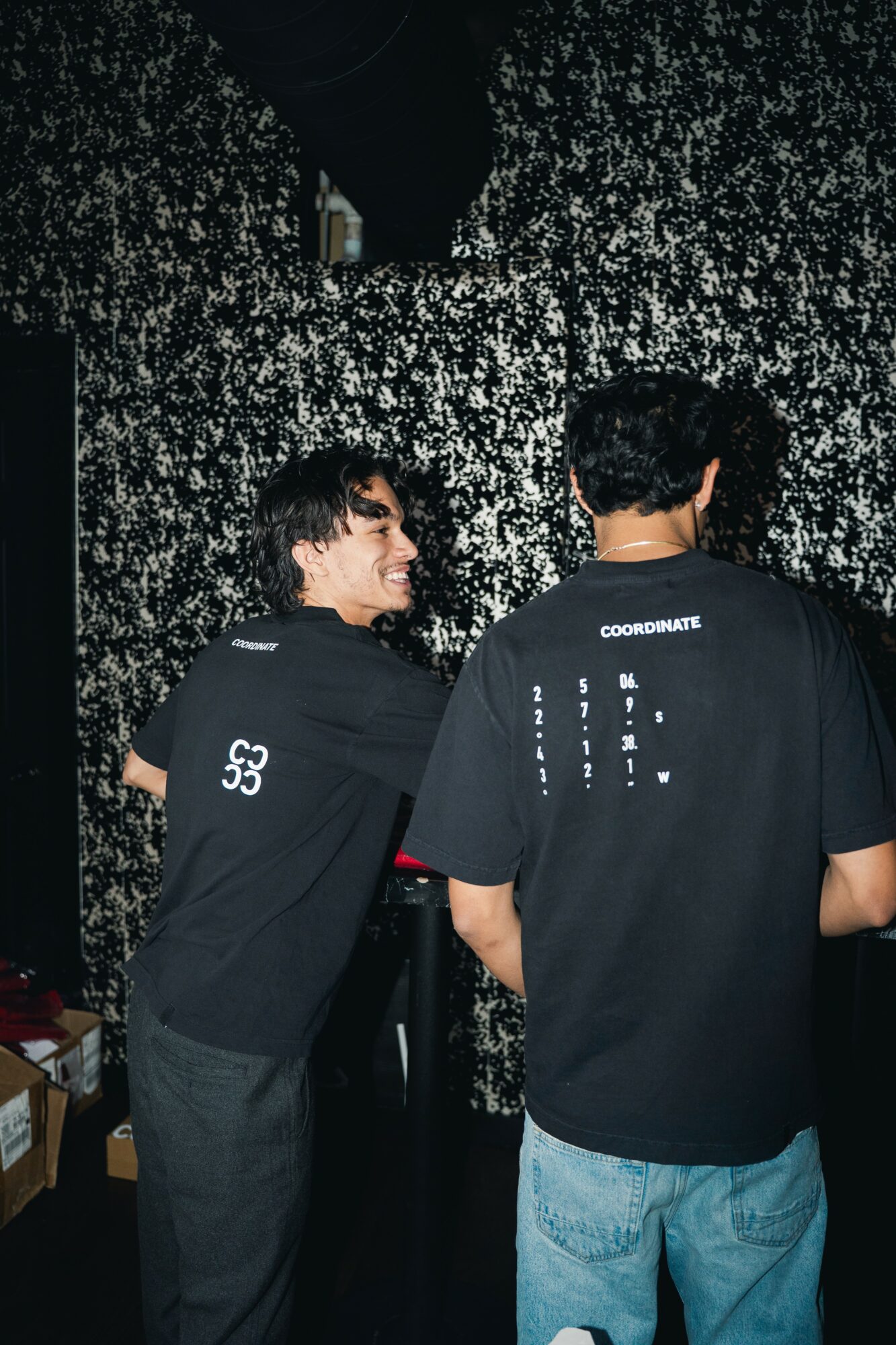Two people with dark hair wearing black shirts stand against a black and white speckled background, facing away from the camera.