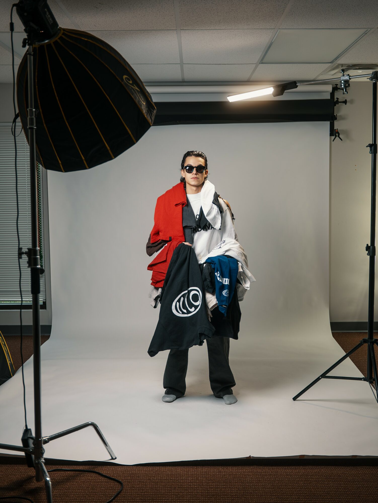Person standing in a photography studio with a white backdrop, wearing sunglasses and a red jacket, holding clothing items.