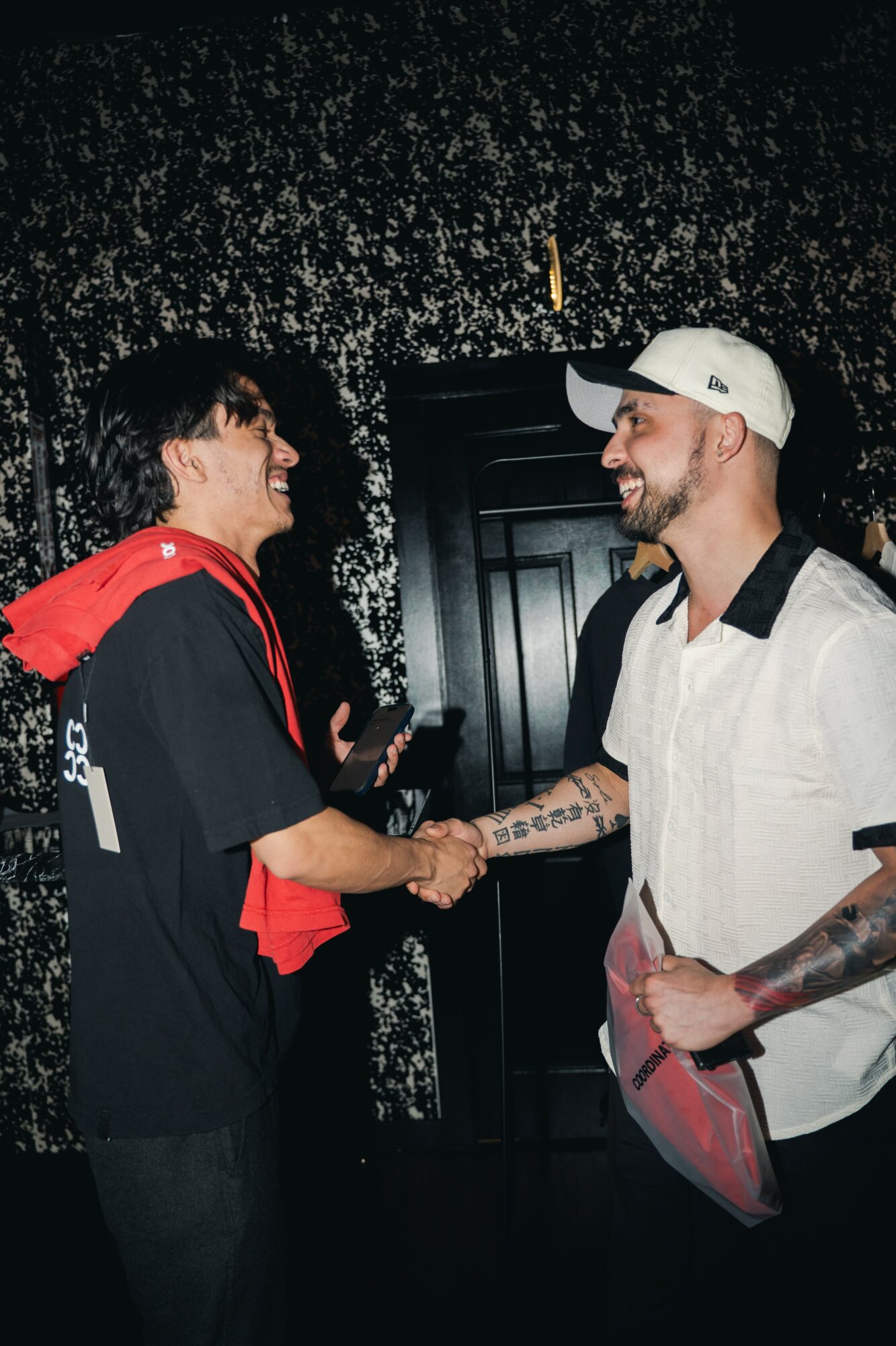 Two people shake hands and smile, one wearing a black and red shirt, the other in a white shirt and cap, in front of a dark background.