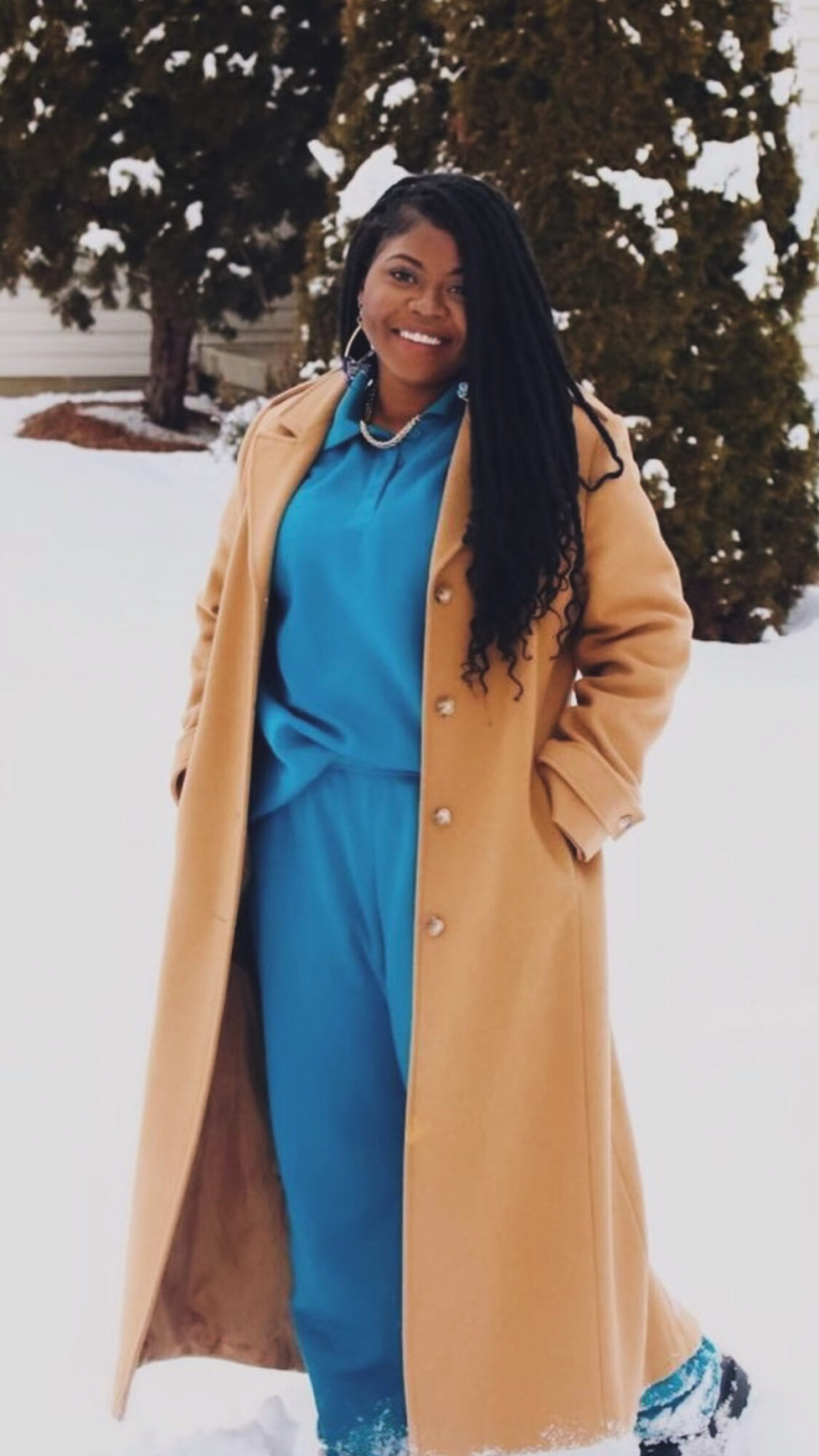 Woman smiling outdoors in winter, wearing a blue outfit and tan coat, with snow and trees in background.