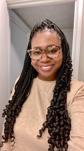 Smiling woman with glasses and long twisted hair, wearing a beige top, indoors.