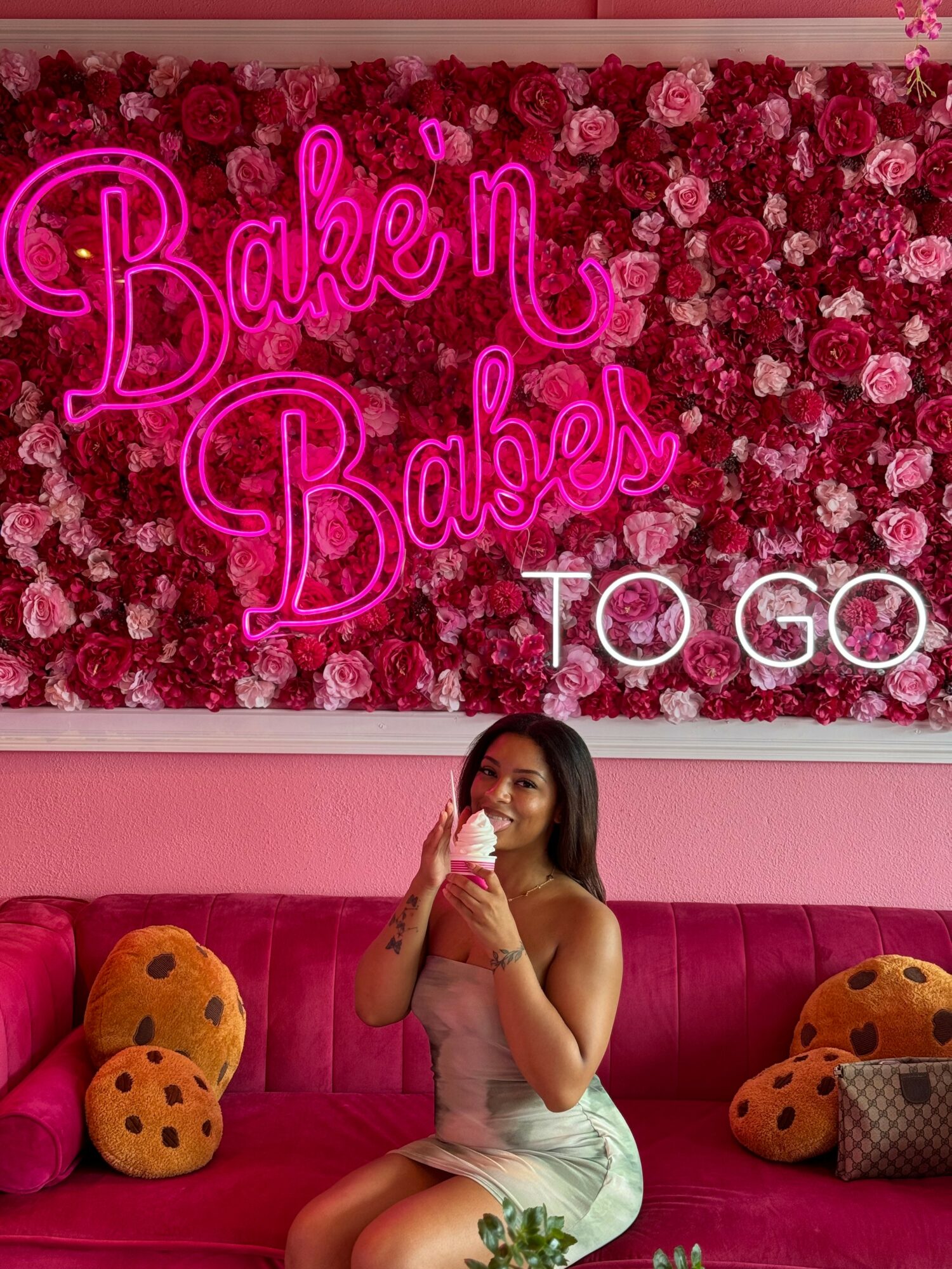 Woman sitting on pink couch with pink and orange plush pillows, holding ice cream, in front of a floral wall with neon sign.