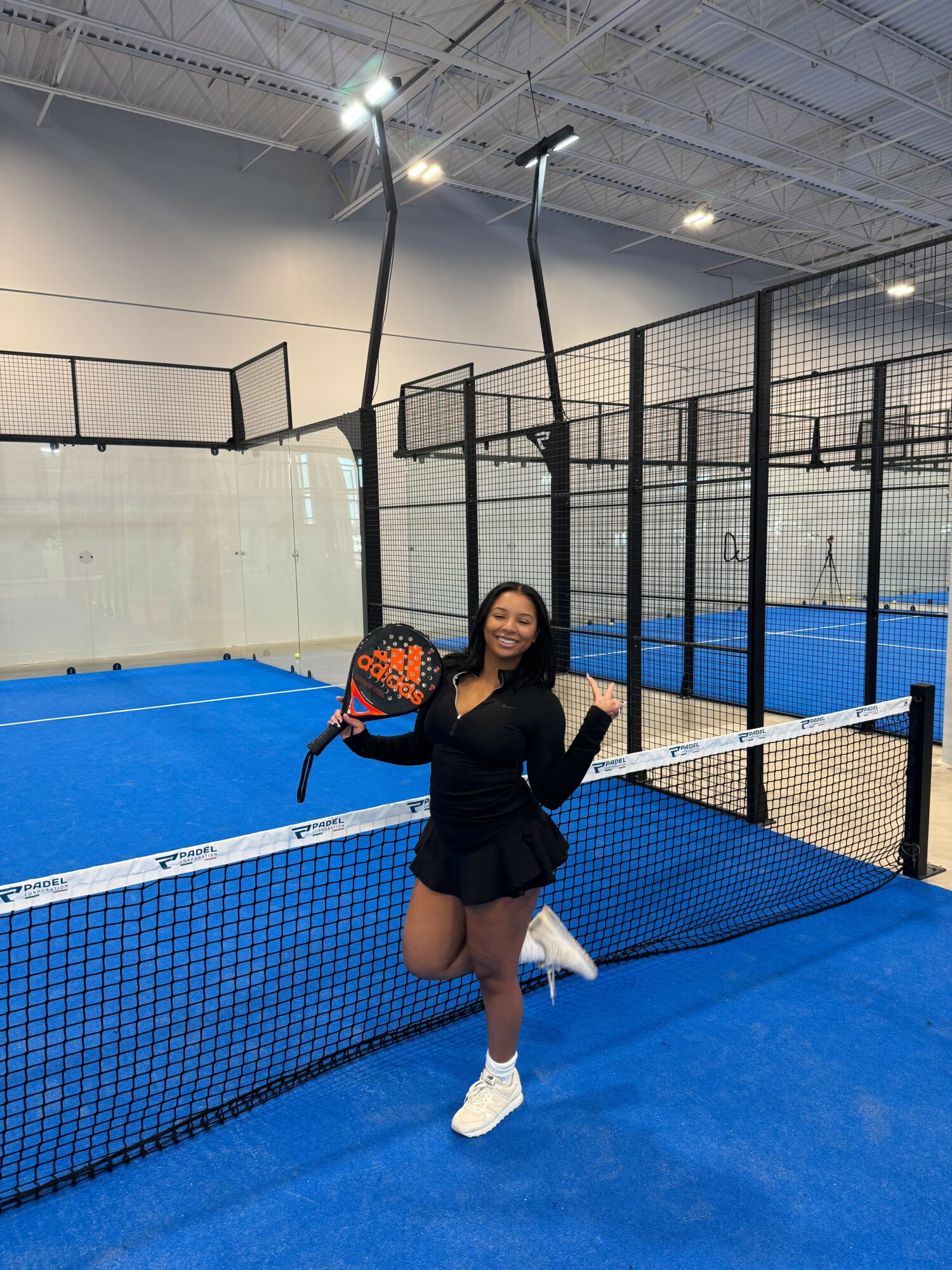 Young woman in black outfit and white sneakers holding a tennis racket, smiling on blue indoor tennis court.
