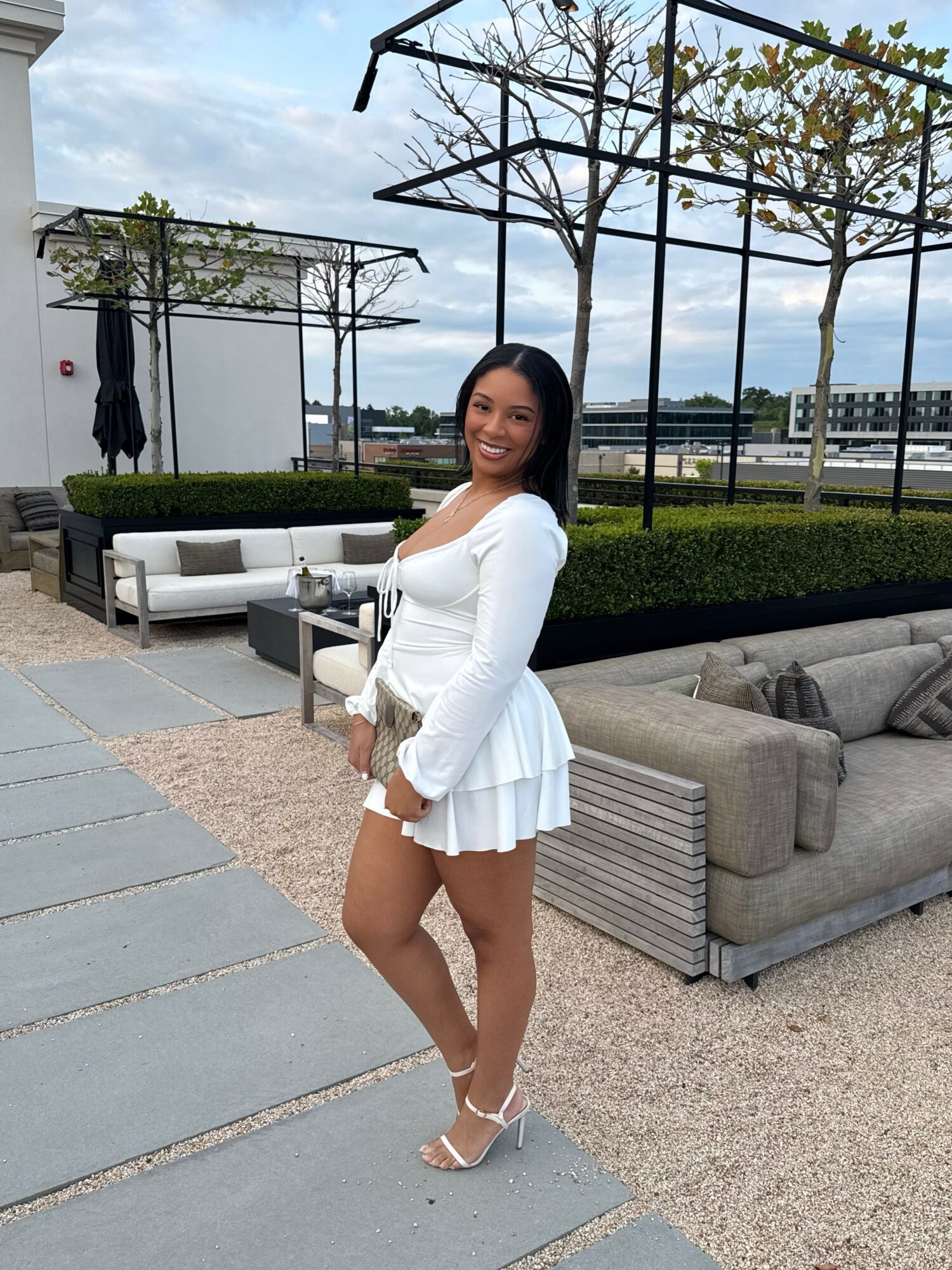 Smiling woman in white dress and heels standing outdoors near seating area with trees and sky in background.