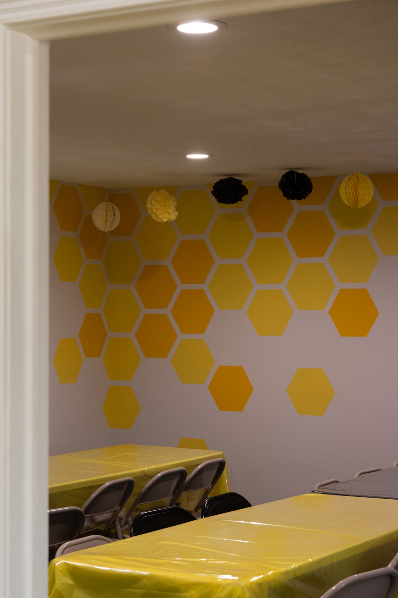 Room with yellow hexagon wall pattern, tables, and chairs, ceiling lights, and black and yellow decorations on ceiling.