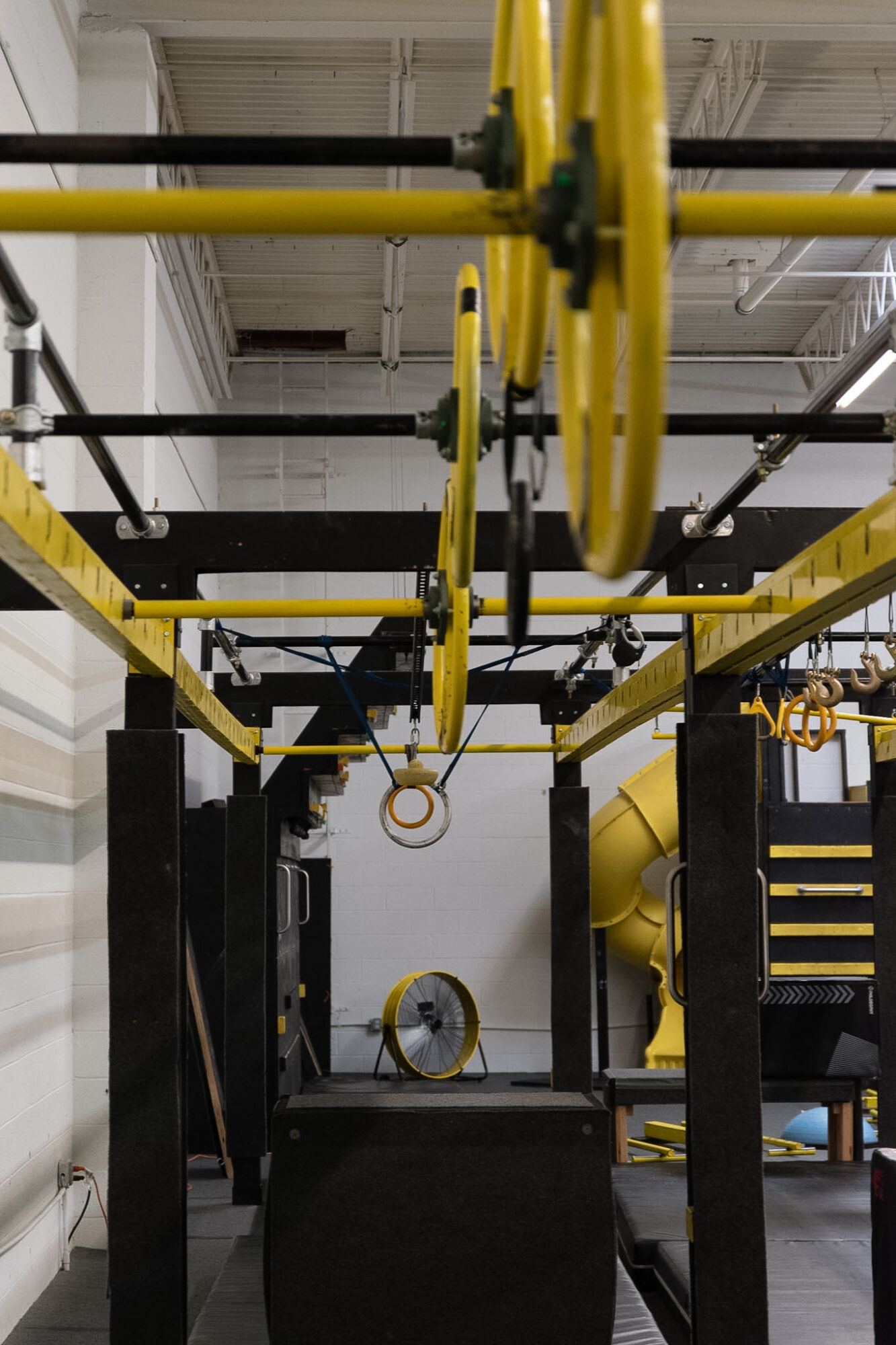Gym equipment with yellow and black structures, gymnastic rings, and a yellow slide in the background.