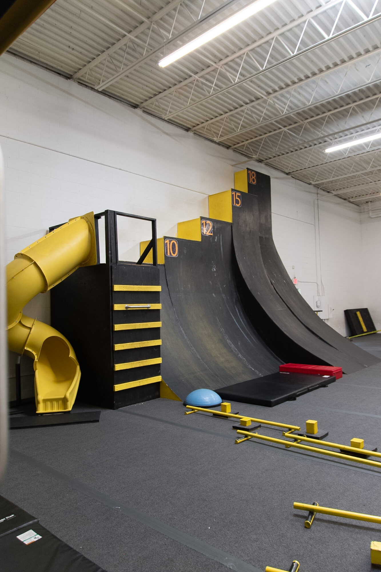 Indoor skate park with a large black ramp, yellow slide, and various equipment on the floor, ceiling with metal beams.