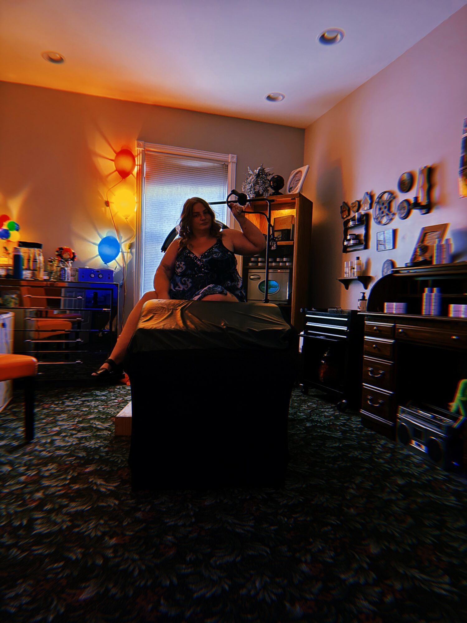 Person sitting on a bed in a dimly lit room with colorful lights, shelves, and wall decorations.