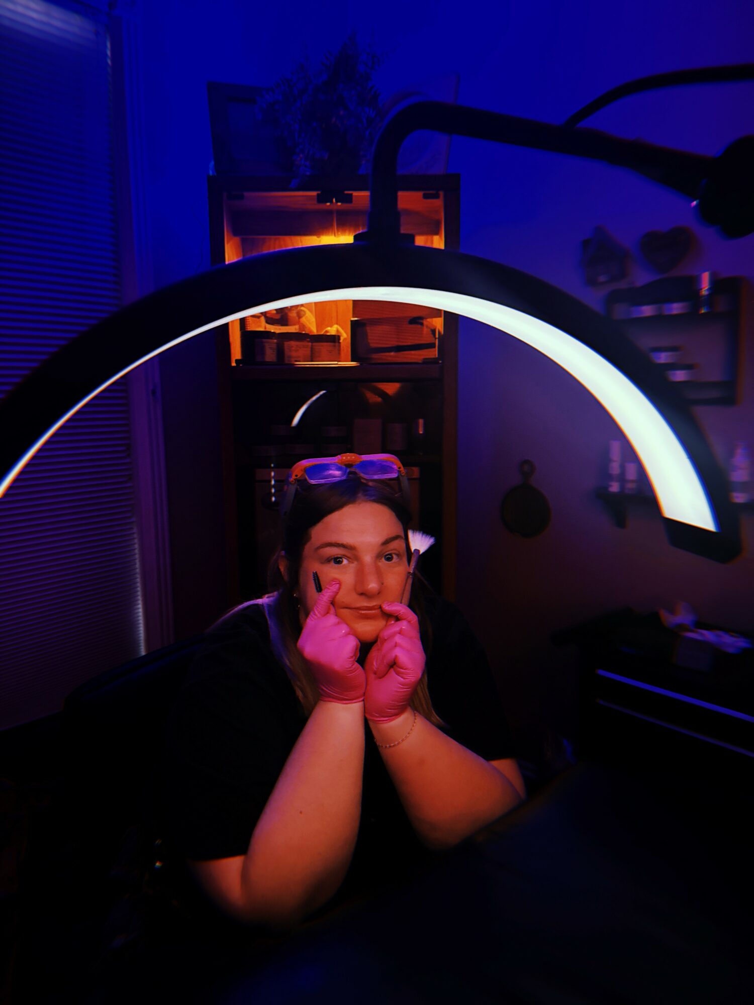 Person with glasses on head, pink gloves, resting chin on hands, illuminated by a curved light, in a dark room.