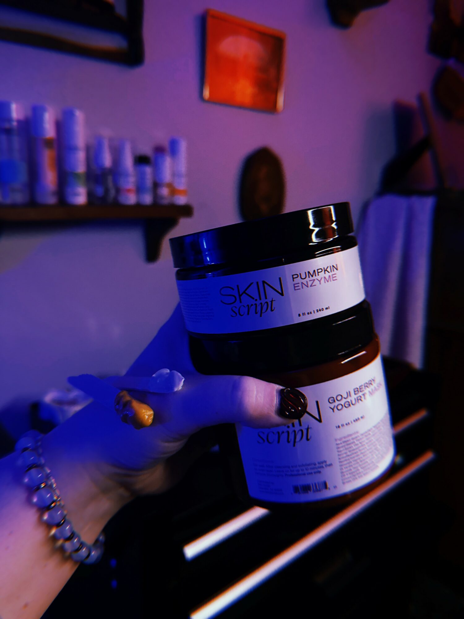 Hand holding two skincare jars in a dimly lit room with wall art and shelves, blue and purple lighting.