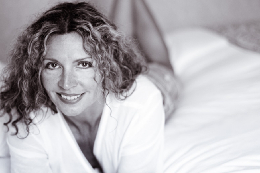 Woman with curly hair smiling, lying on bed, in a white shirt, in a bedroom setting.