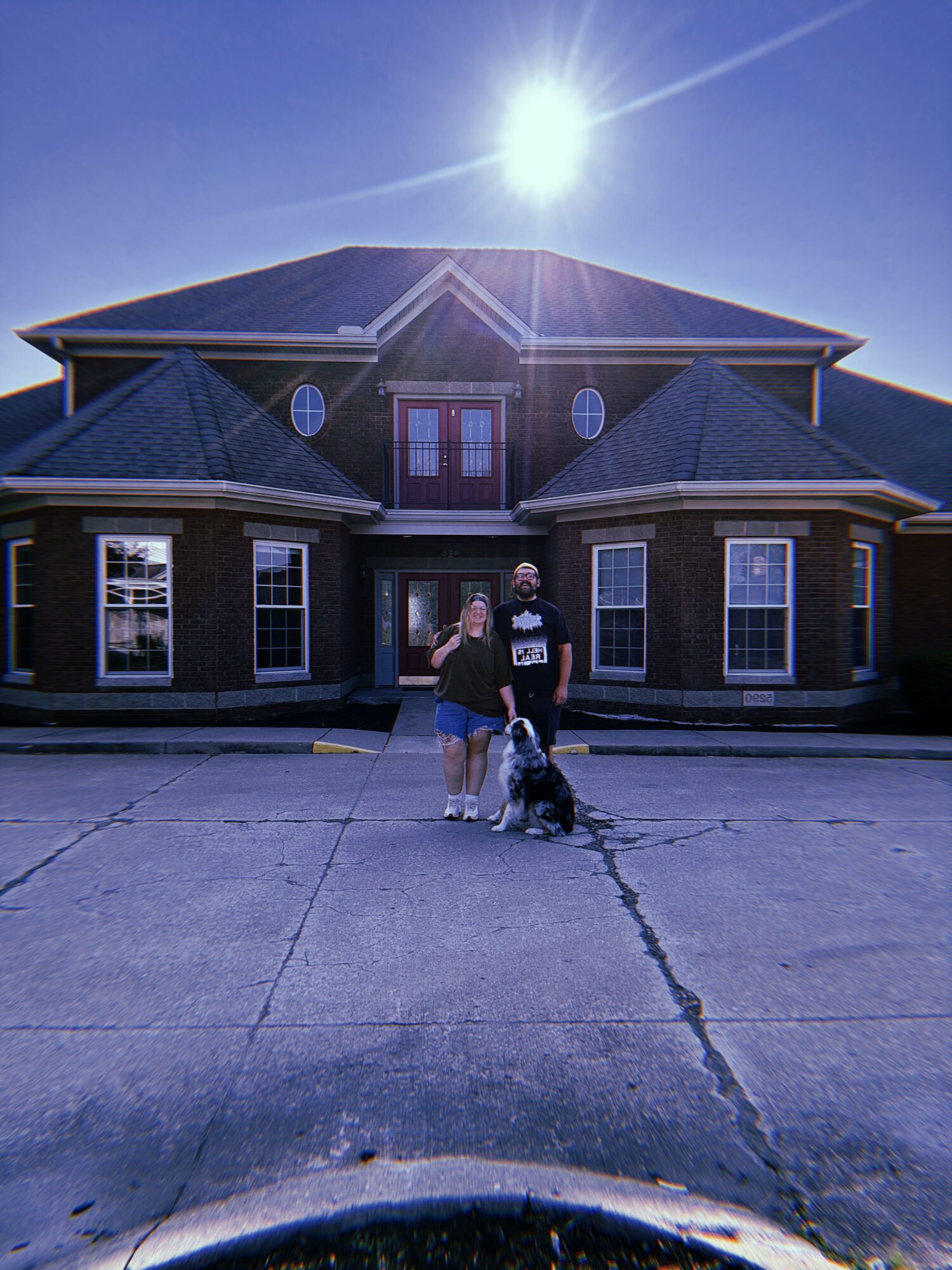 Two people and a dog standing in front of a large house with a bright sun overhead, clear sky, and parking lot.