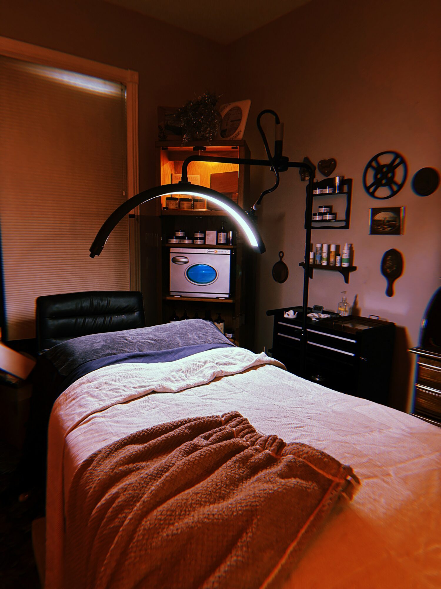 Bedroom with bed, black headboard, and wall-mounted lamp, shelves with bottles, and a small cabinet, warm lighting.