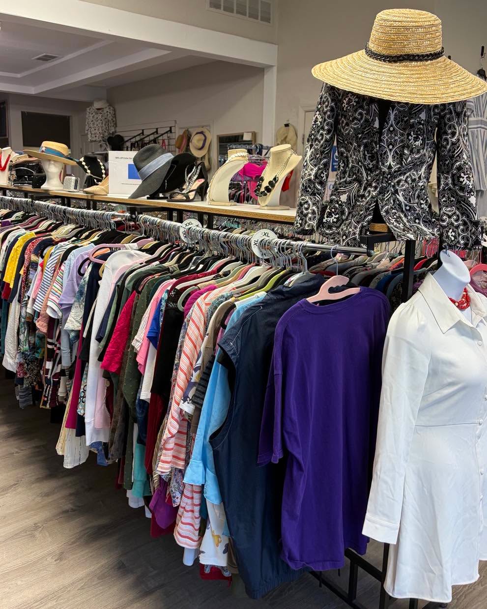 Clothing rack with various colorful shirts, dresses, and a white coat, with hats displayed on mannequins and shelves in the background.