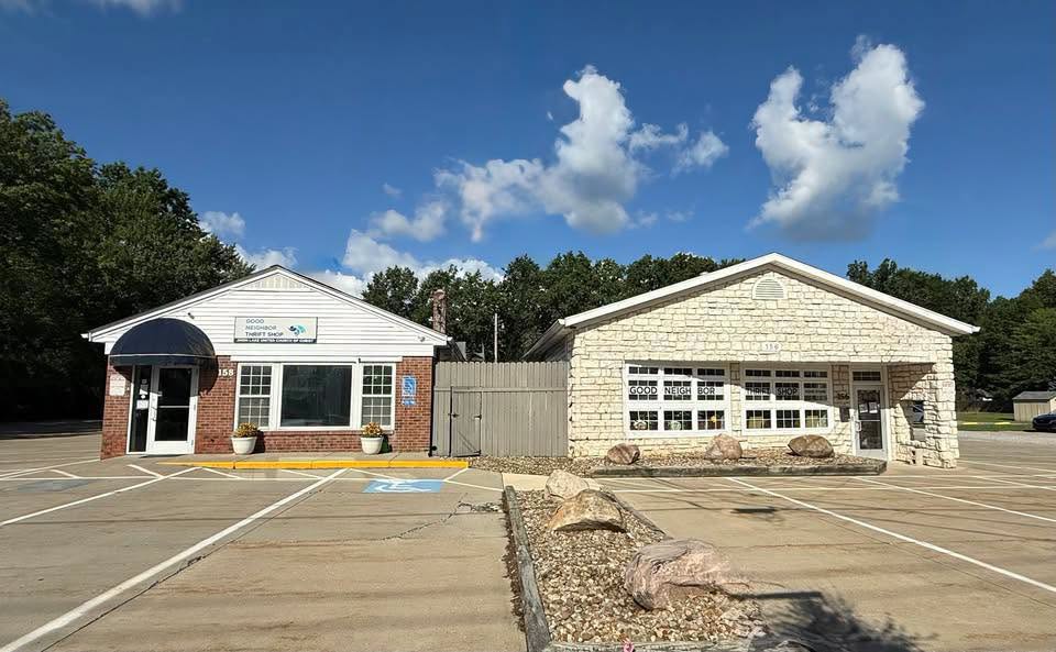 Two small buildings with parking lot in front, blue sky with clouds, trees in background.