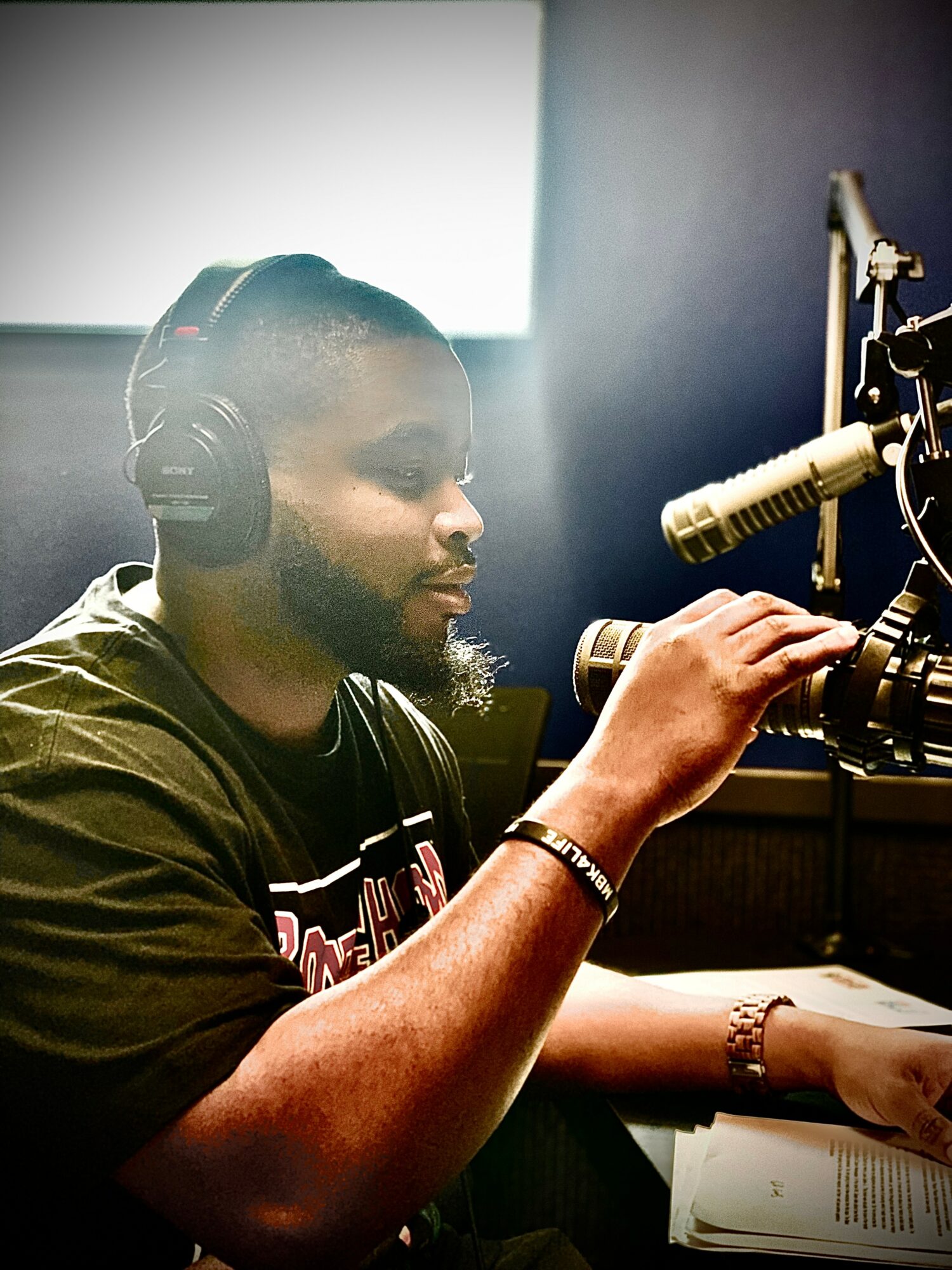 Person wearing headphones speaking into a microphone in a recording studio, with a window behind them.